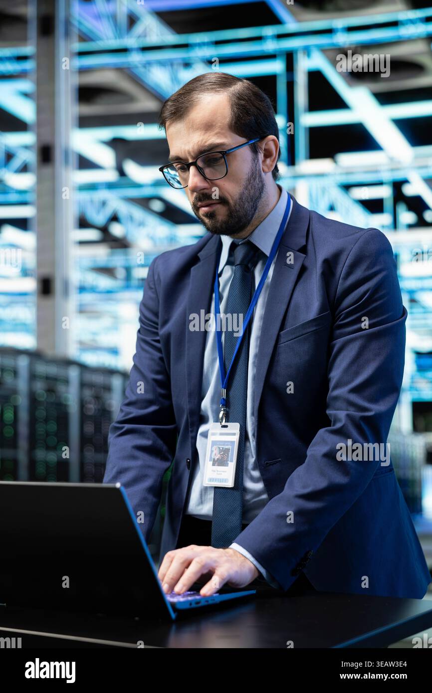 Data center programmer at desk configuring server settings with laptop software. Worker in modern high tech server farm using notebook to do maintenance tasks on IT infrastructure machines Stock Photo