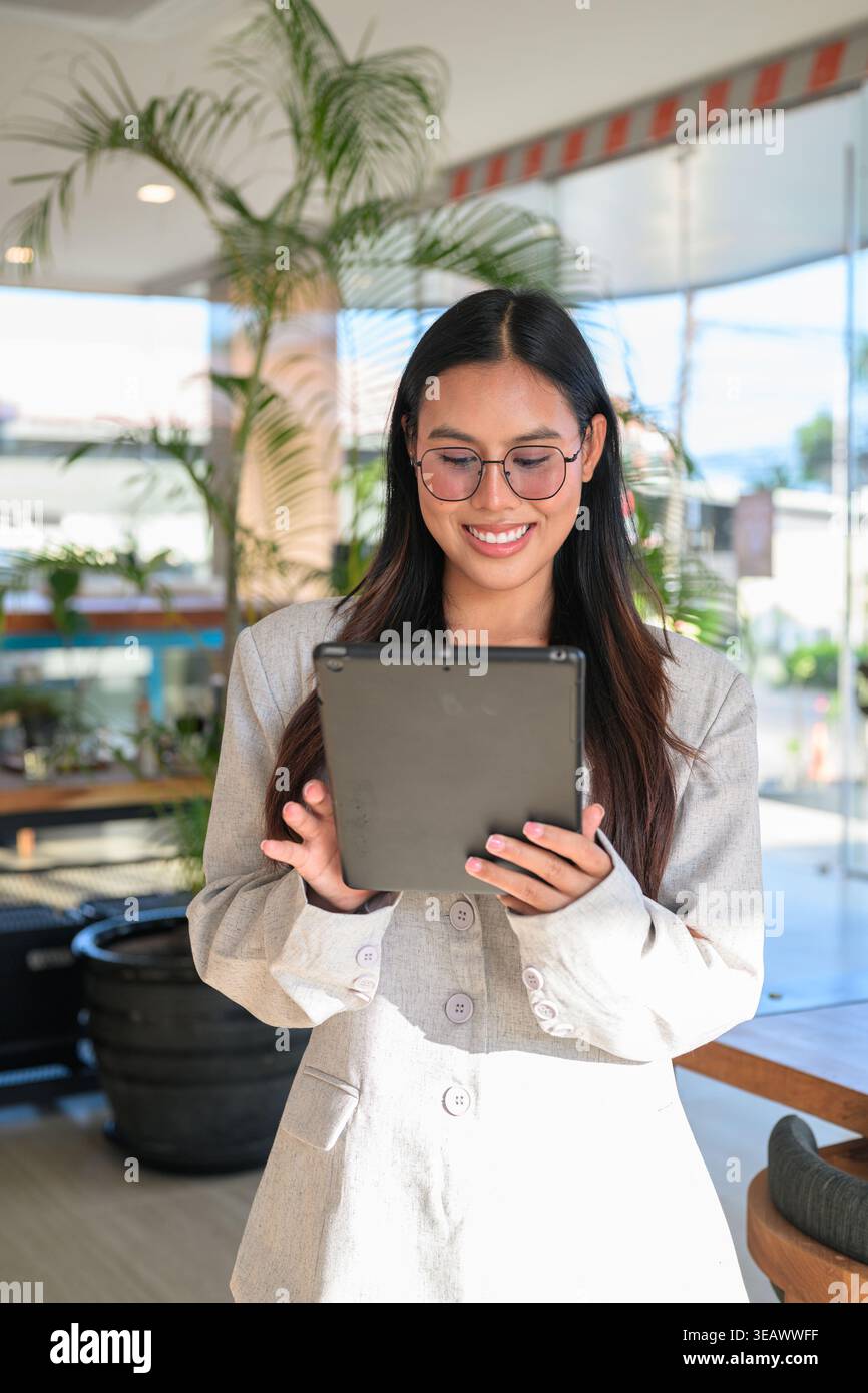 Young Asian woman from the Isan region of Thailand, showcasing a natural smile and honest expression while using a digital tablet in a contemporary ca Stock Photo