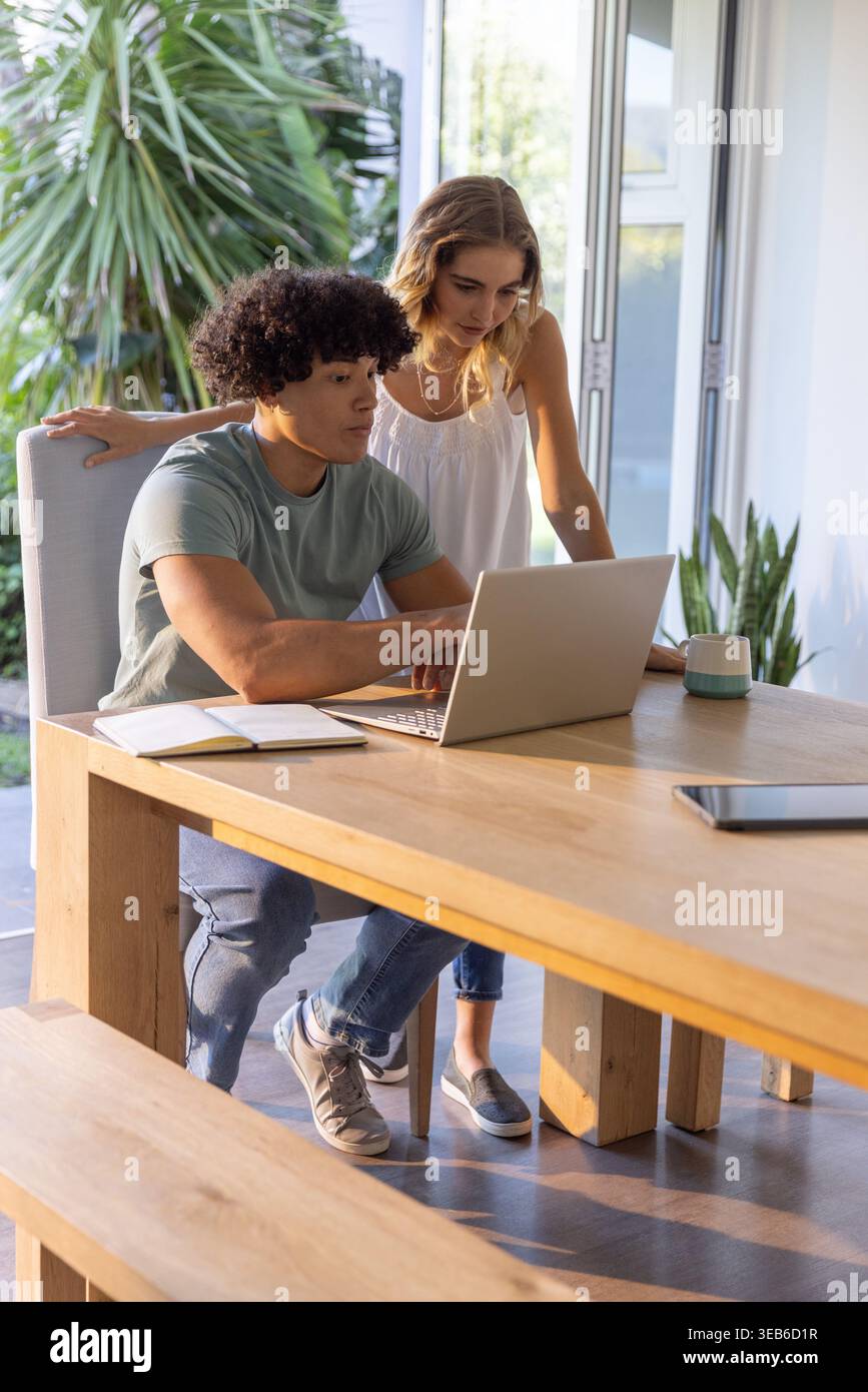 Diverse couple working at wooden dining table using silver laptop while being surrounded by plants Stock Photo