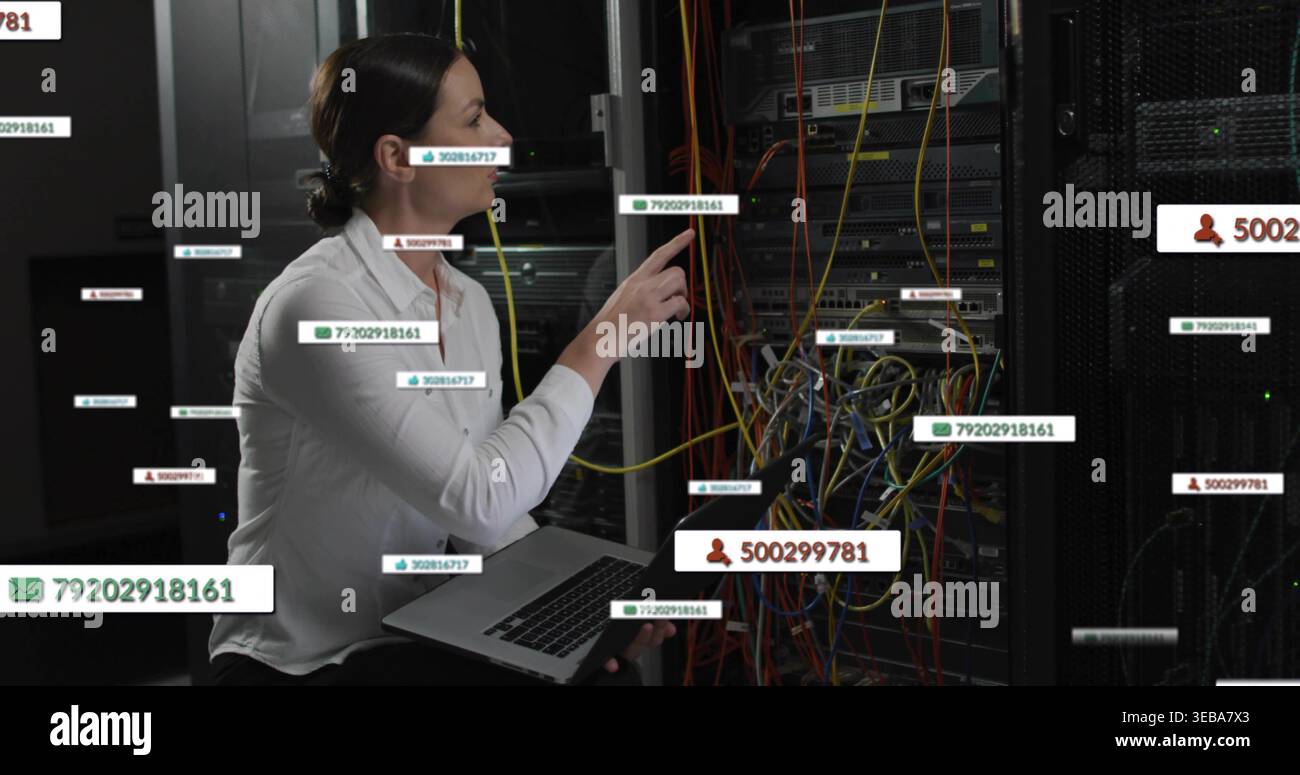 Kneeling IT tech in white blouse touching patch panel with laptop in datacenter, showing cables Stock Photo