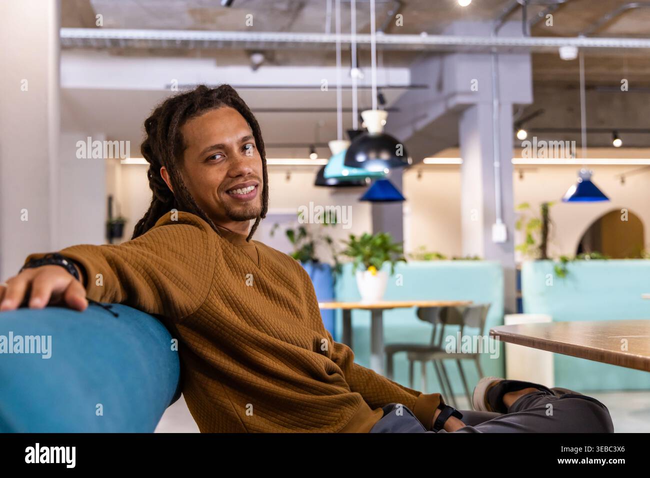 African American man in late twenties sitting on blue bench in coworking lounge checking wristwatch Stock Photo
