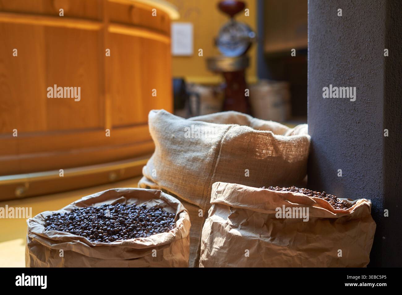 Close-up of dark roasted coffee beans contained in open burlap and brown paper sacks resting on a wooden floor beside a structural column. The Stock Photo