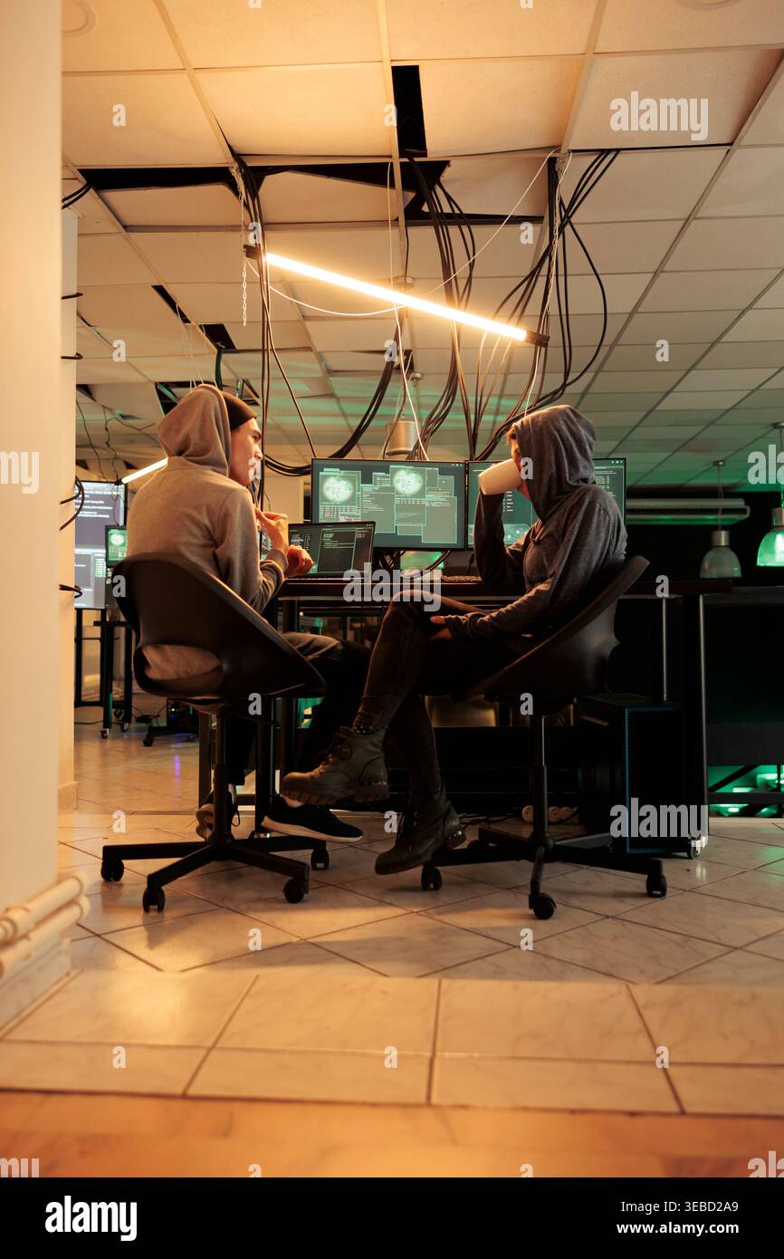 Young hackers coding trojan virus program to hack system, IT specialists programming illegal firewall encryption. Two cybercriminals developing malicious software late at night. Stock Photo