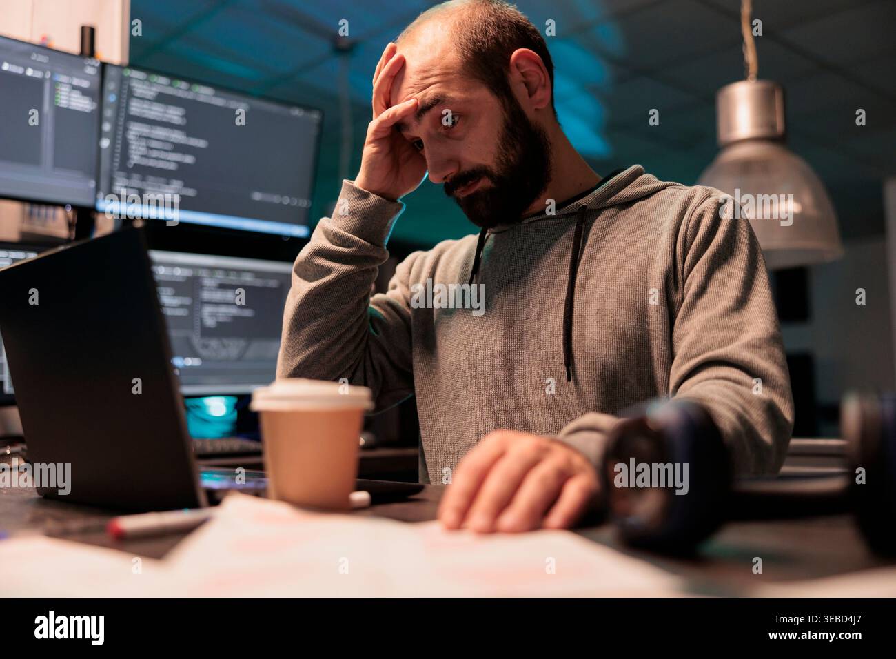 Stressed overworked developer programming html code on laptop and multiple monitors, working under stress after hours. Male app programmer feeling frustrated and tired coding data script. Stock Photo
