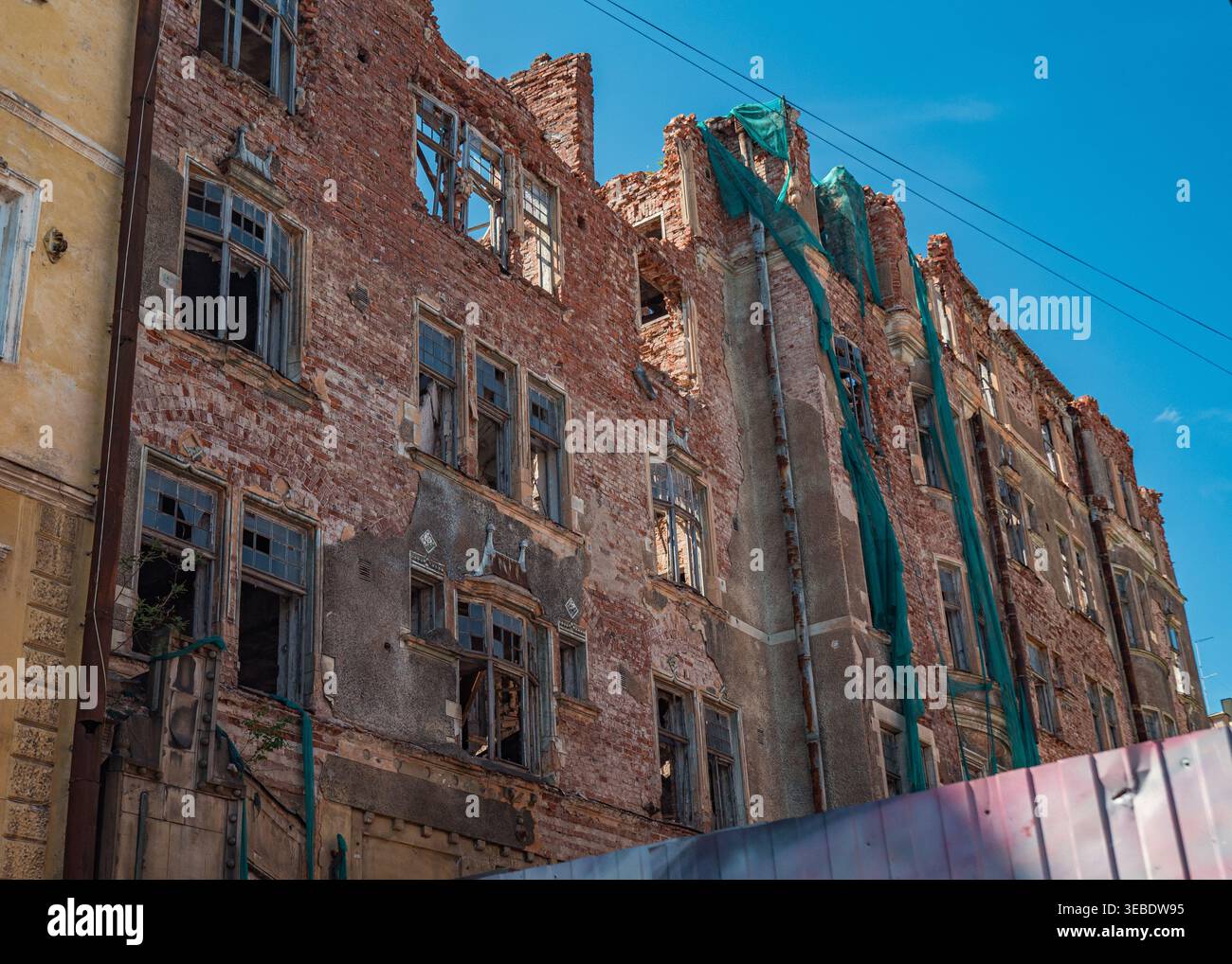 Damaged urban brick building with shattered windows and crumbling walls featuring green safety netting under blue sky Stock Photo