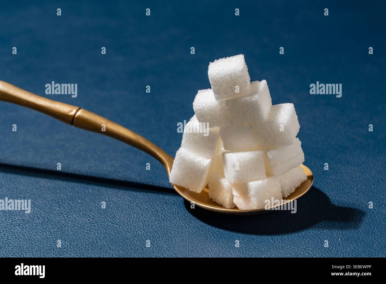 Pile of sugar cubes in spoon on blue background. High stack of white refined sugar cubes in golden spoon on blue textured surface. Concept of sugar ad Stock Photo