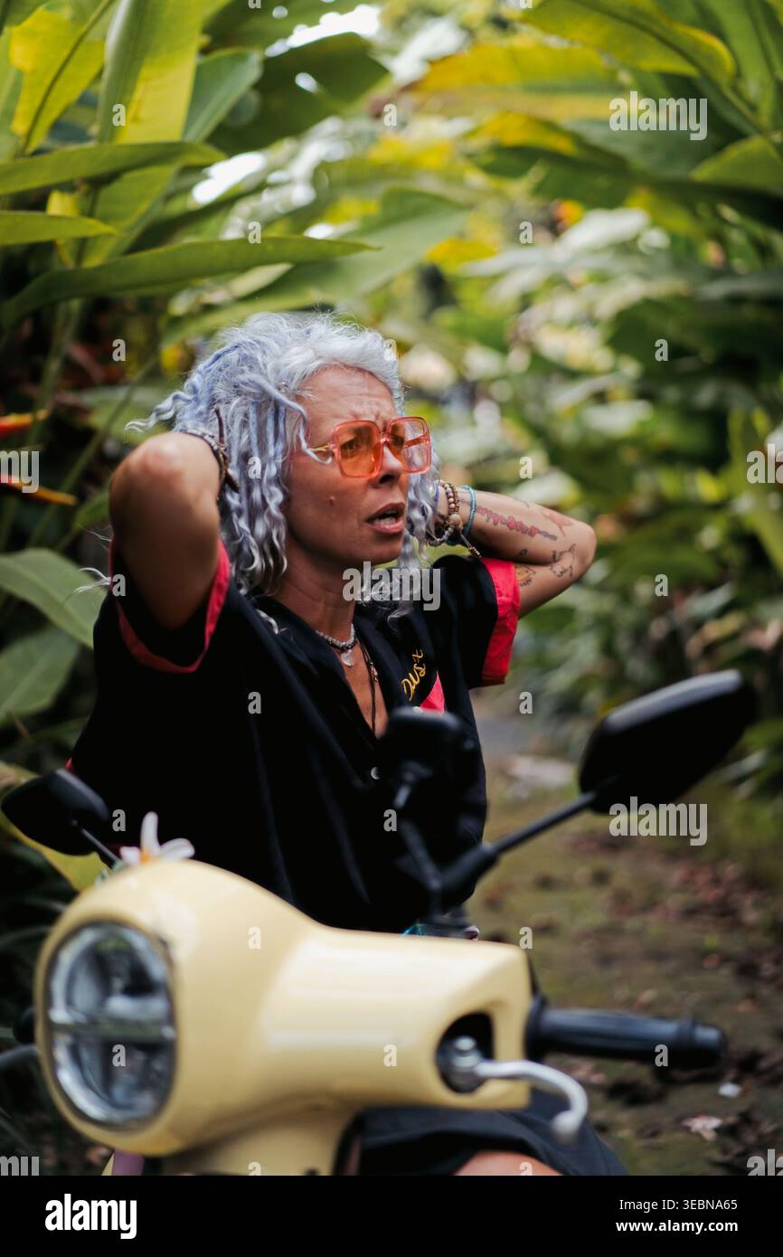 Mature woman on scooter adjusting hair on tropical road, Canggu, Bali Stock Photo