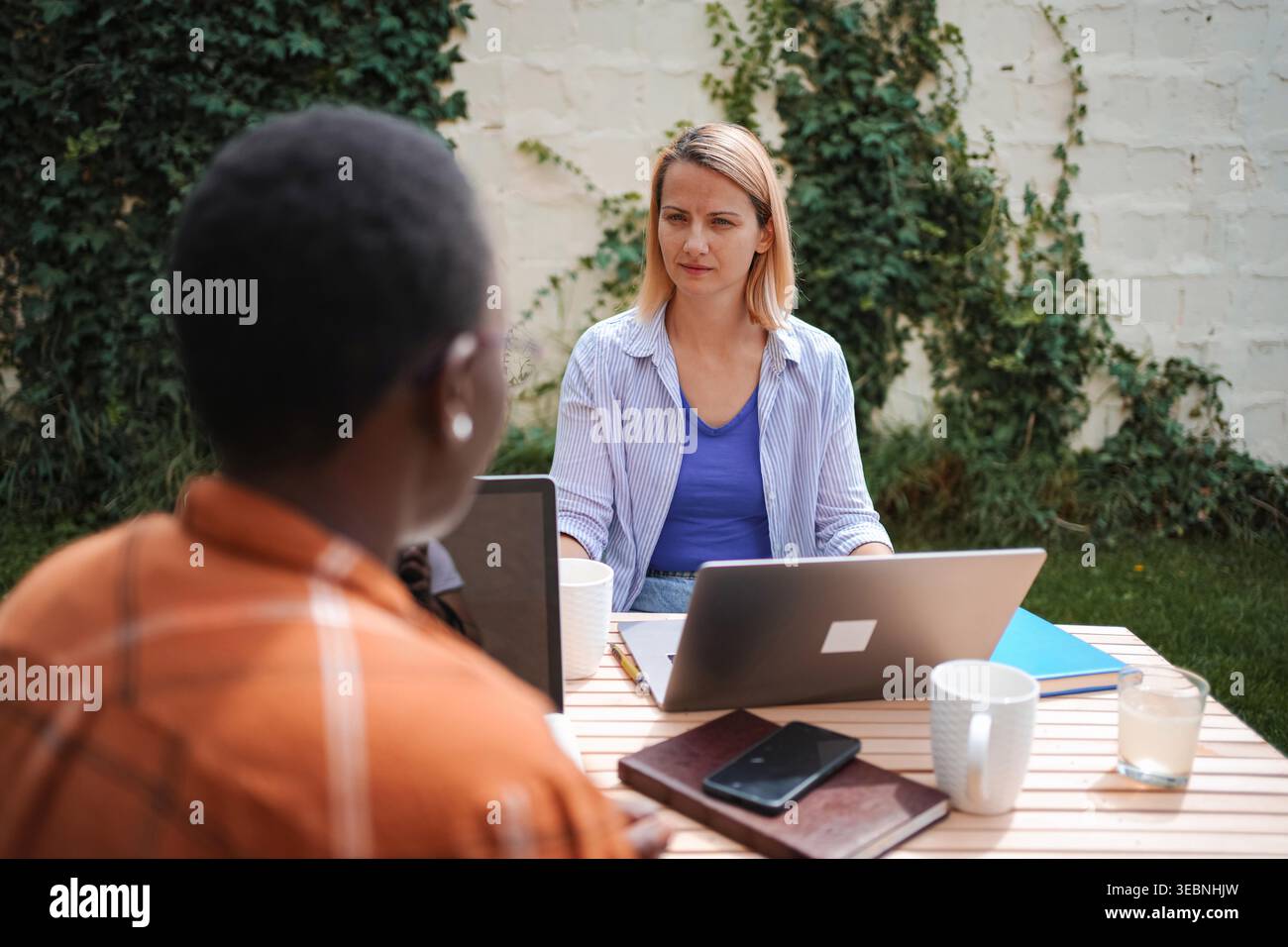 Two businesswomen working remotely outdoors, using laptops and discussing a project Stock Photo