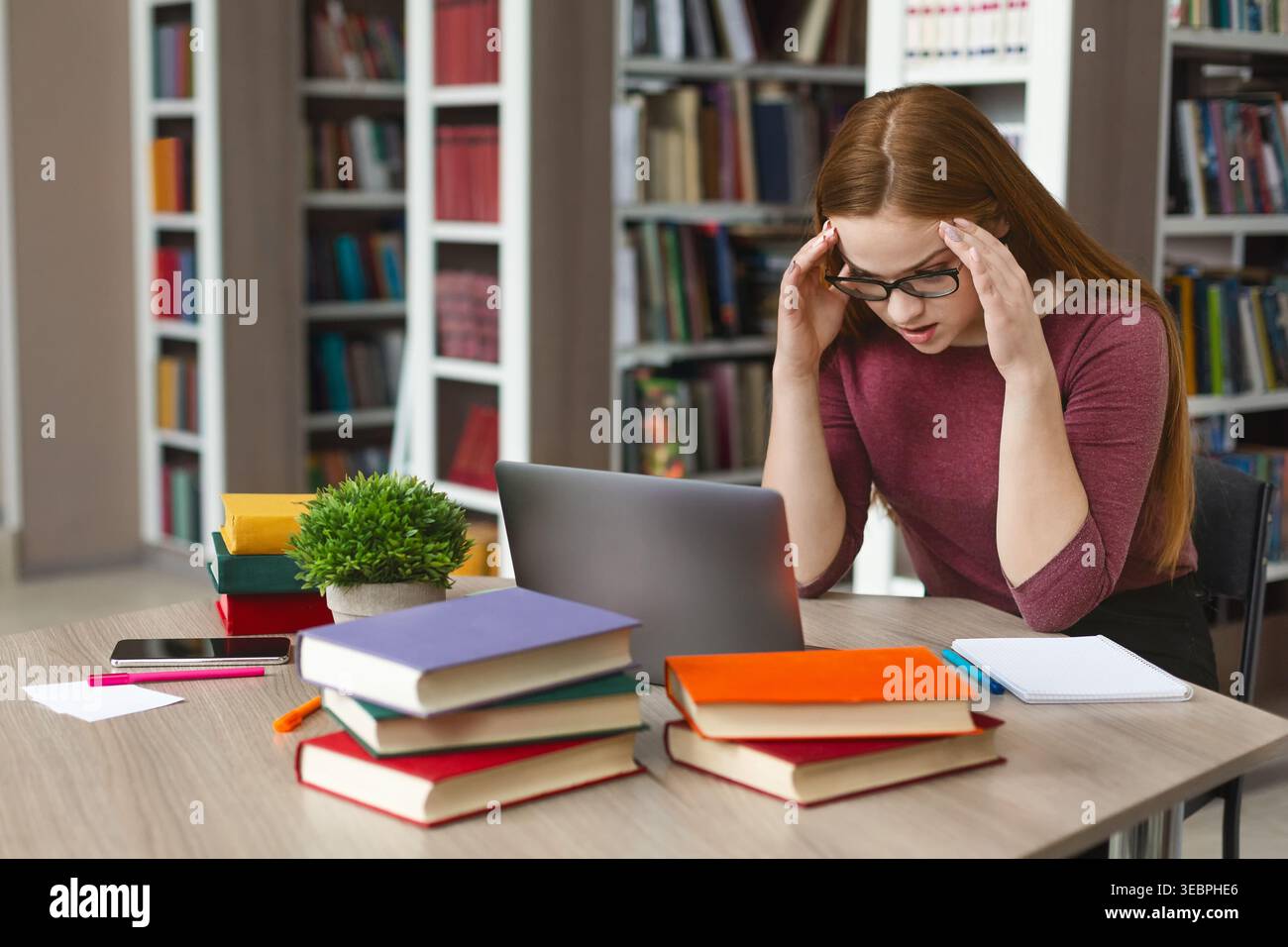 Student Studying With Stress in Library Surrounded by Books During Study Session Stock Photo