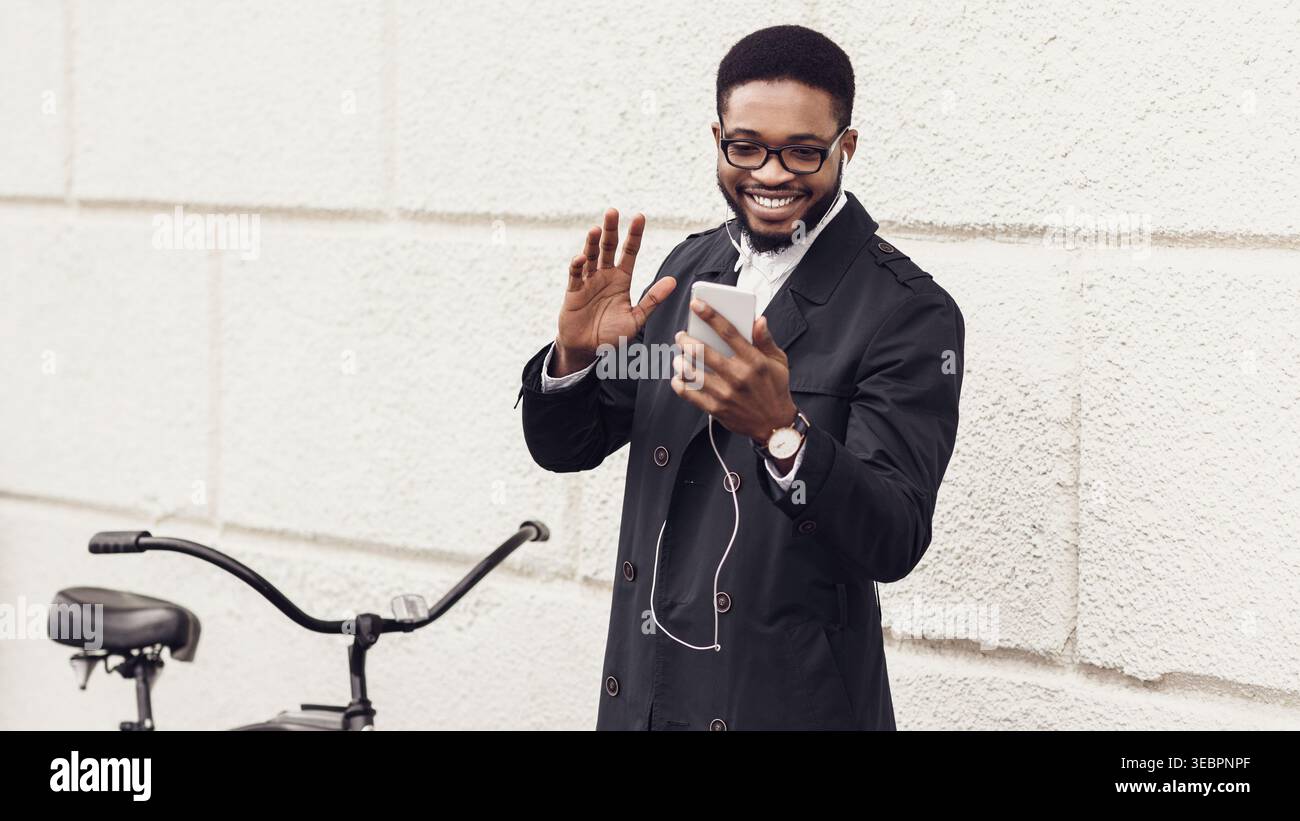 Man Smiling and Waving While Using Smartphone in Urban Setting With Bicycle Nearby Stock Photo
