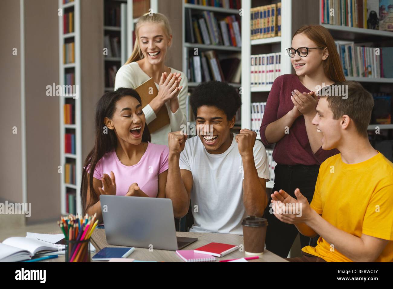 Group of Students Celebrating Success While Using a Laptop in a Bright Library Setting Stock Photo