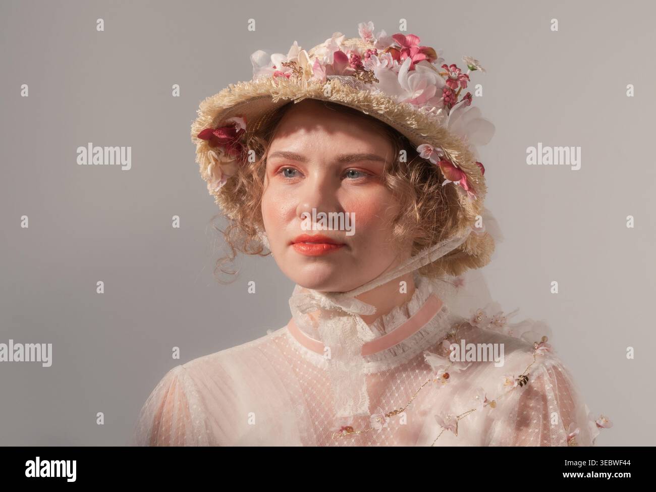 Young woman models elaborate flower hat and vintage-inspired sheer top in a bright studio Stock Photo