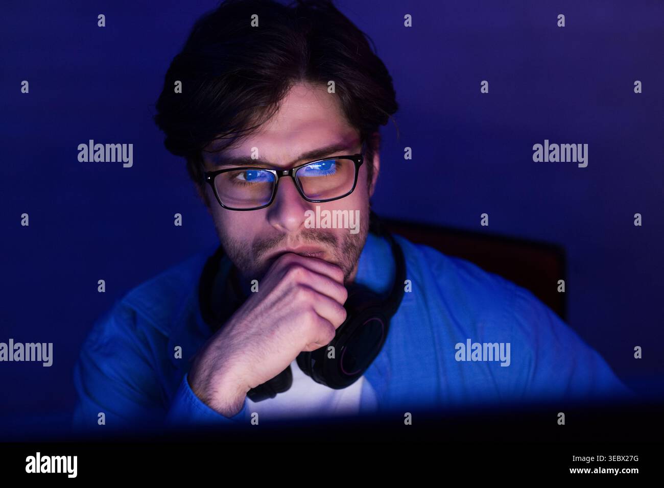 Young Man Wearing Glasses Deep in Thought While Working at Computer in Dimly Lit Room Stock Photo