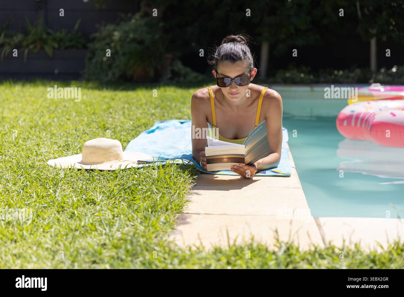 Mid-adult woman lying on blue towel reading paperback at backyard pool, wearing yellow swimsuit Stock Photo