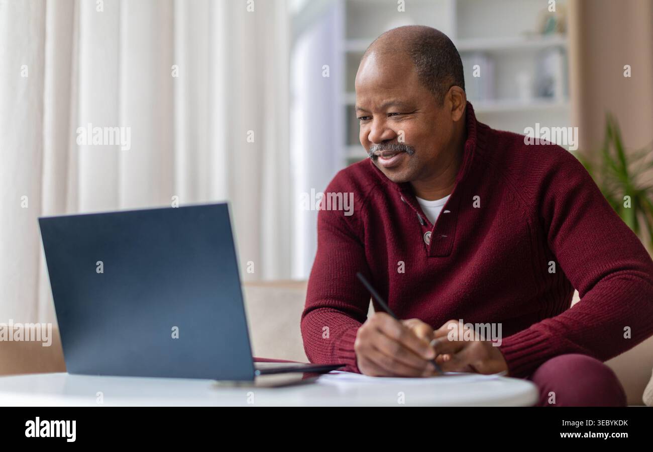 Smiling mature black man taking notes while working on laptop at home Stock Photo