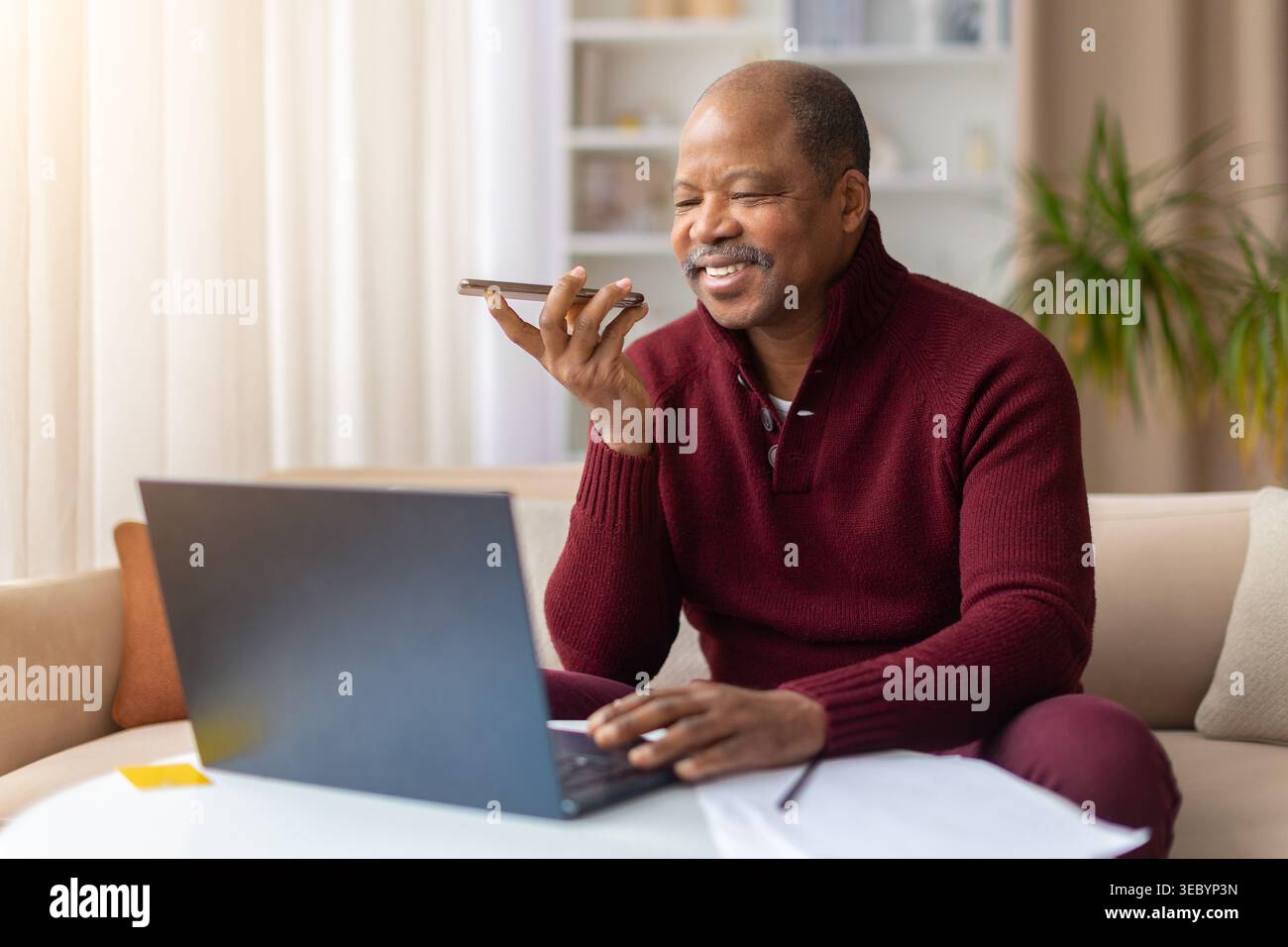 Black man using smartphone voice assistant while working on laptop at home Stock Photo