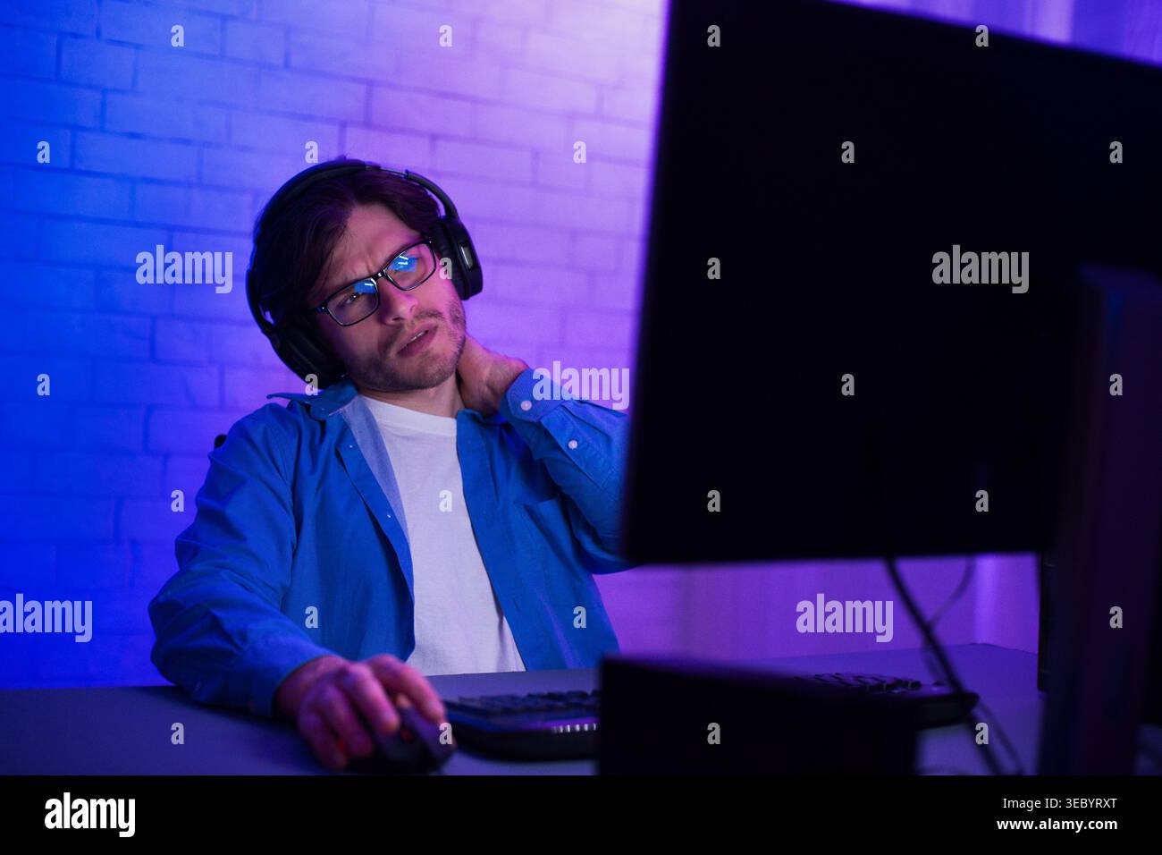 Young Man Using Computer While Wearing Headphones and Looking Thoughtfully at the Screen Stock Photo