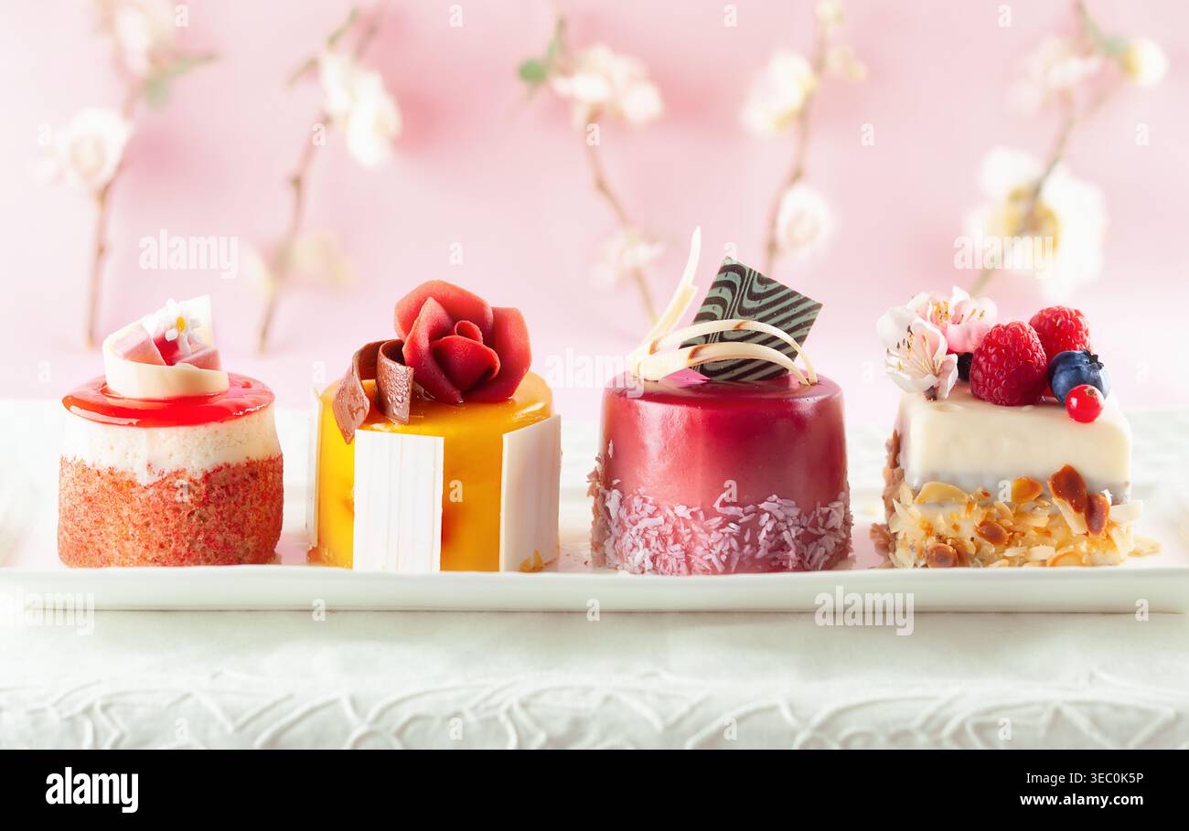Various mini cakes on a white plate. Sweets decorated with fresh berries and flowers for holiday. Stock Photo
