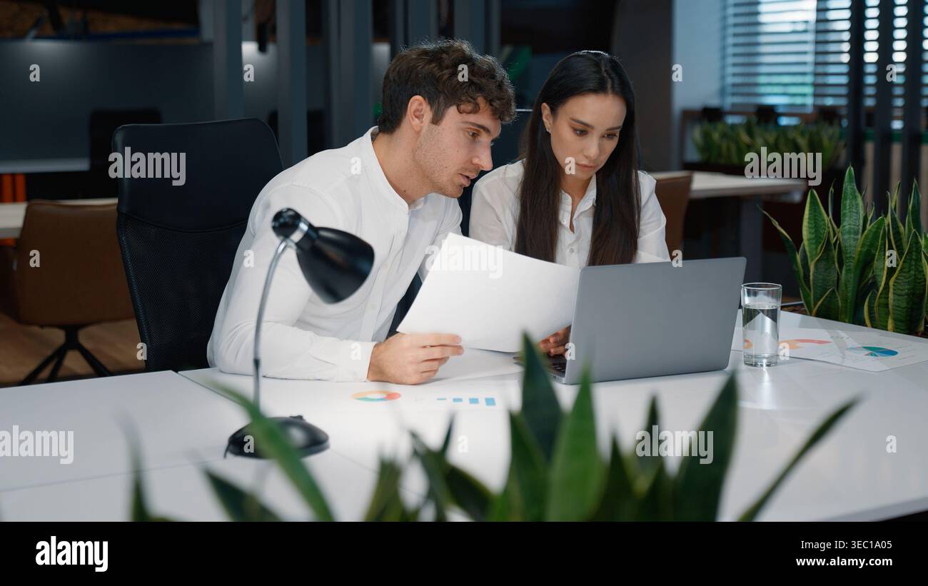 Two business partners talking looking in papers with stats data graphs of project marketing result reading documents computer work in office Stock Photo