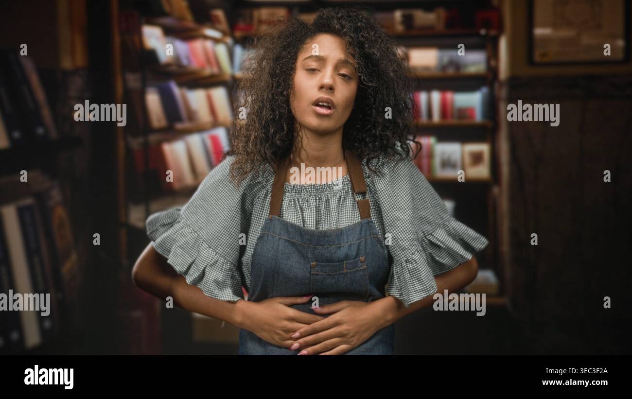 Teenage woman with curly hair wearing denim apron clutches stomach with ...