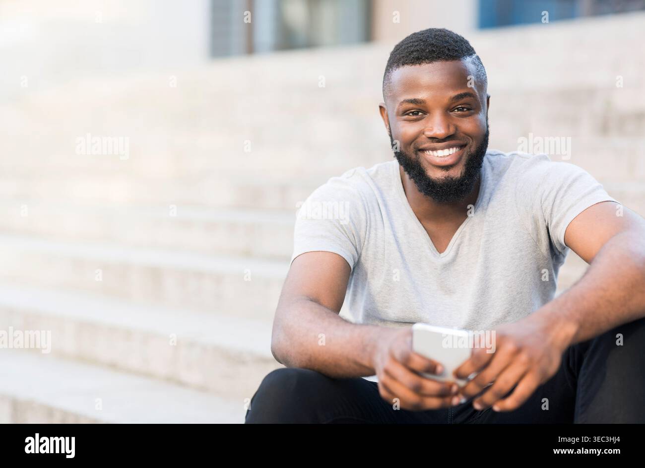 Young Man Smiling While Sitting on Steps and Using Smartphone Outdoors in Bright Sunlight Stock Photo