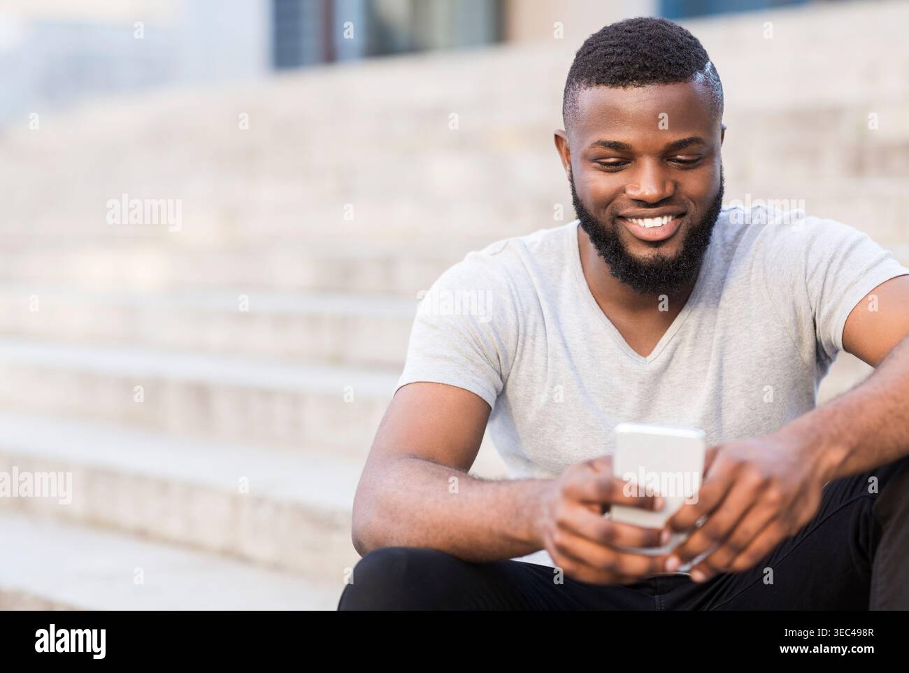 Young Man Sitting on Steps Using Smartphone Outdoors While Smiling in a Casual Setting Stock Photo