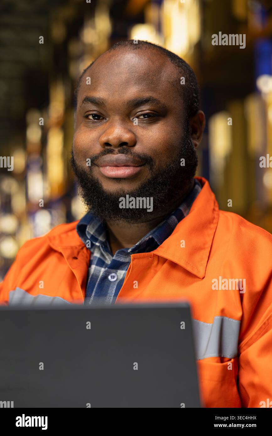 Portrait of black man using laptop for supply chain management, coordinating import export shipment and parcel tracking for e-commerce operations. Manual labor for large business facility. Stock Photo