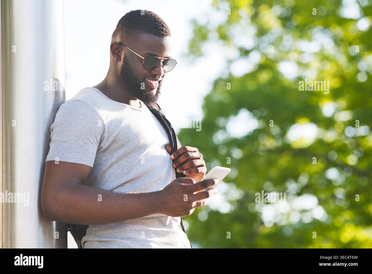 Young Man Using Smartphone While Leaning Against Wall in Bright Outdoor Setting Stock Photo