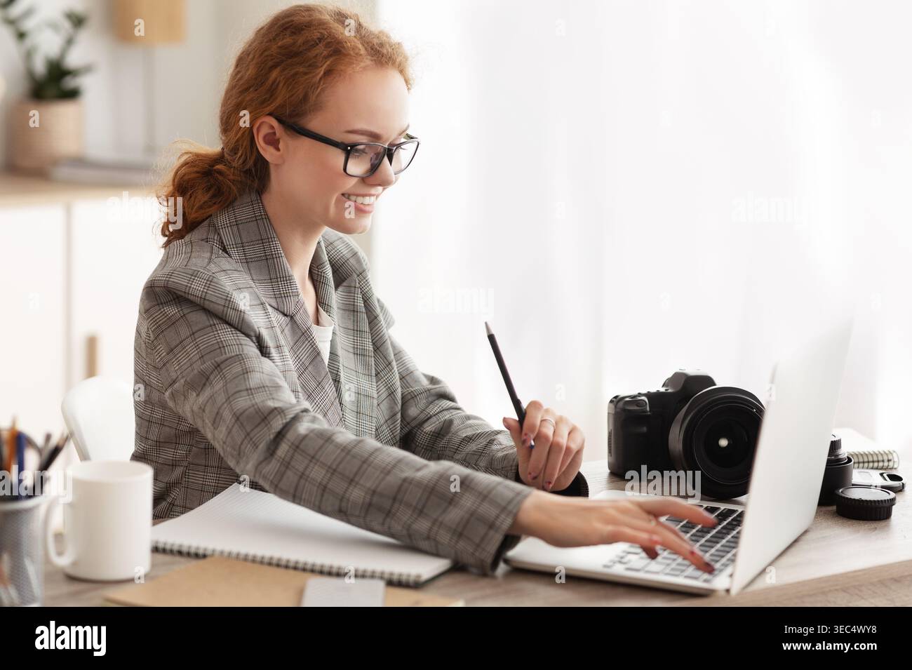 Young Woman Working on Laptop While Taking Notes in Stylish Workspace With Camera Stock Photo