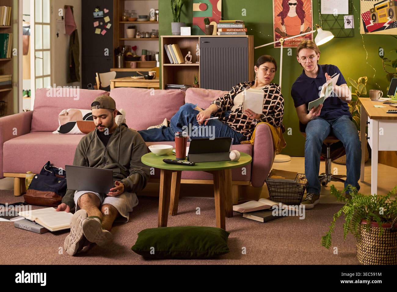 Wide shot of diverse students studying in shared apartment, using laptop and books while discussing coursework. Ideal for education marketing, student housing, campus life content Stock Photo