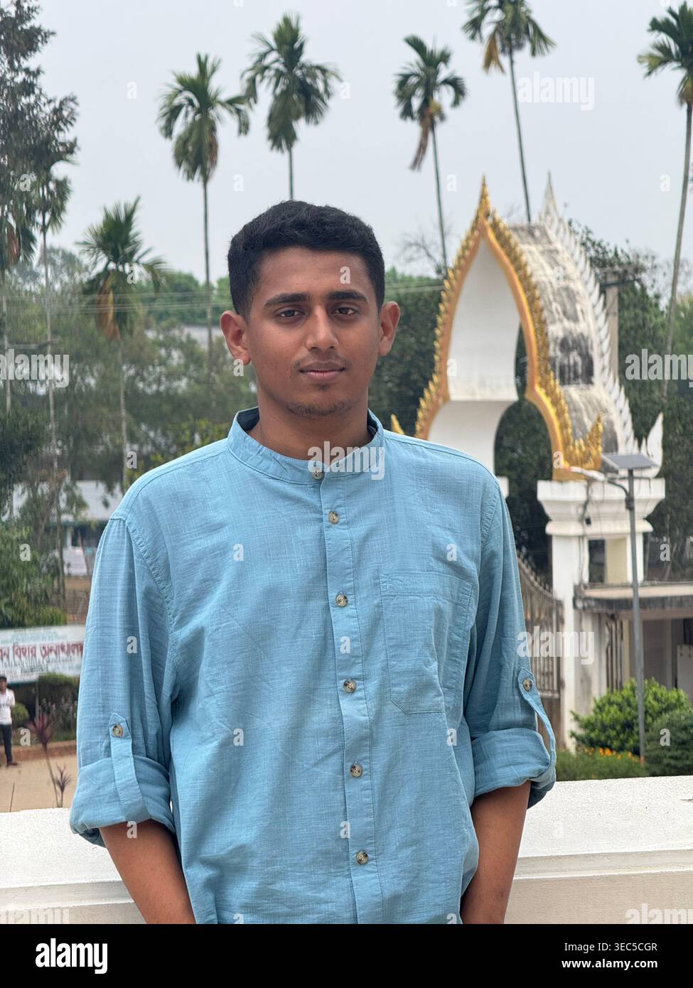 A stylish young man stands confidently at a scenic temple entrance, surrounded by tall palm trees and a peaceful natural backdrop. Wearing sunglasses - Smartphone Captured Stock Image