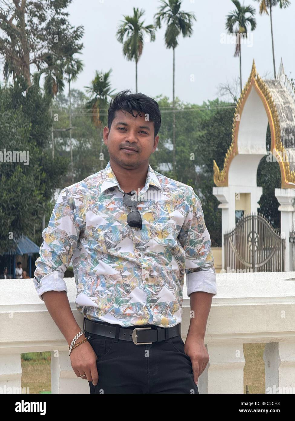 A stylish young man stands confidently at a scenic temple entrance, surrounded by tall palm trees and a peaceful natural backdrop. Wearing sunglasses - Smartphone Captured Stock Image