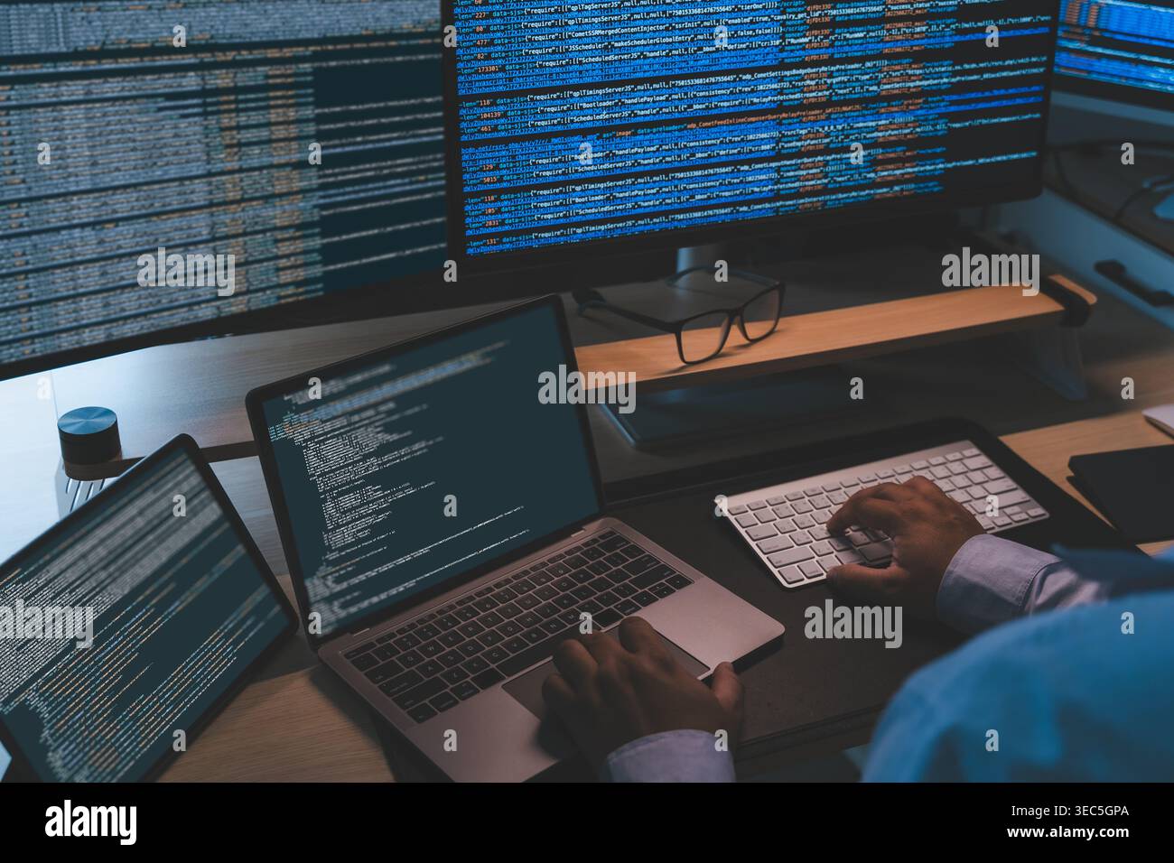 IT programmer working at a multi screen workstation with laptop and monitors displaying source code, showing software development workflow, coding pro Stock Photo