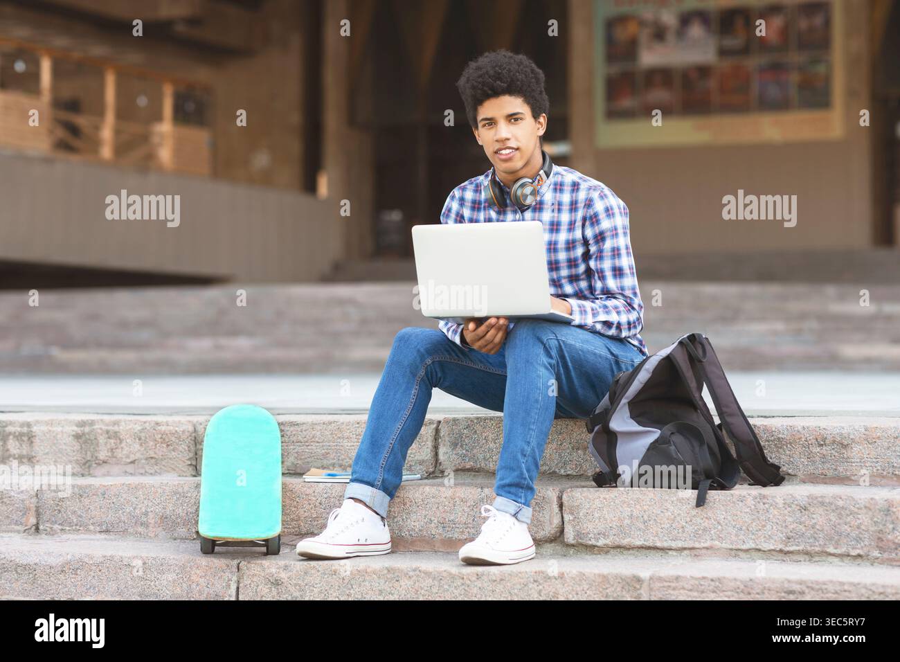 Young Man Using Laptop While Sitting on Steps With Backpack and Skateboard Nearby Stock Photo