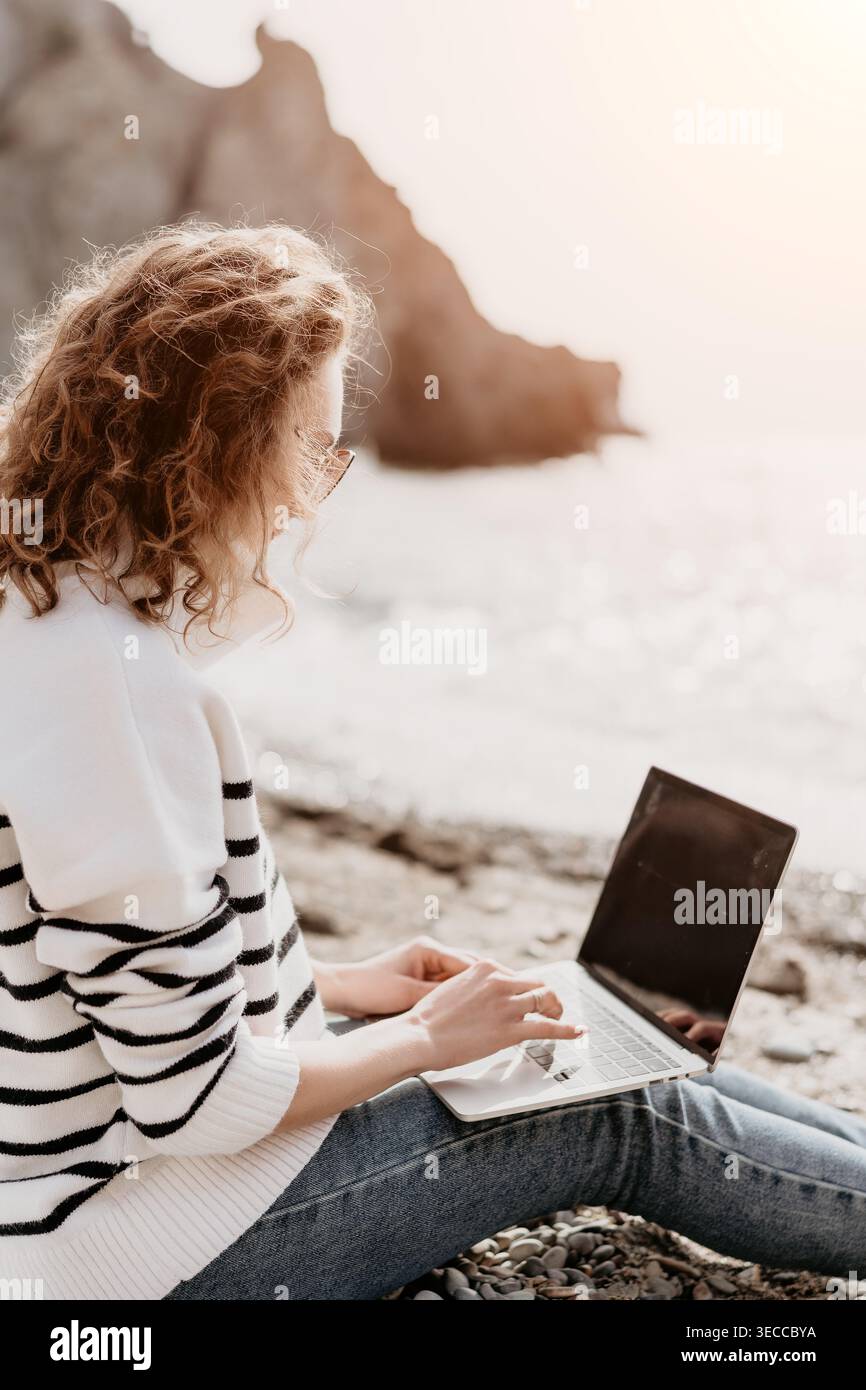Laptop remote beach woman sits on pebbles typing on her computer with scenic ocean and cliffs in the background Stock Photo