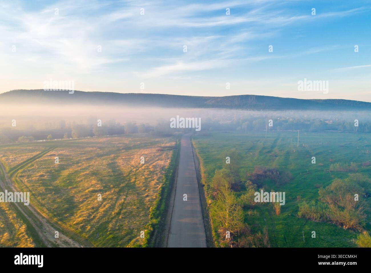 Aerial view captures long country road through sunlit fields, morning mist filling the valley, distant hills under blue sky Stock Photo