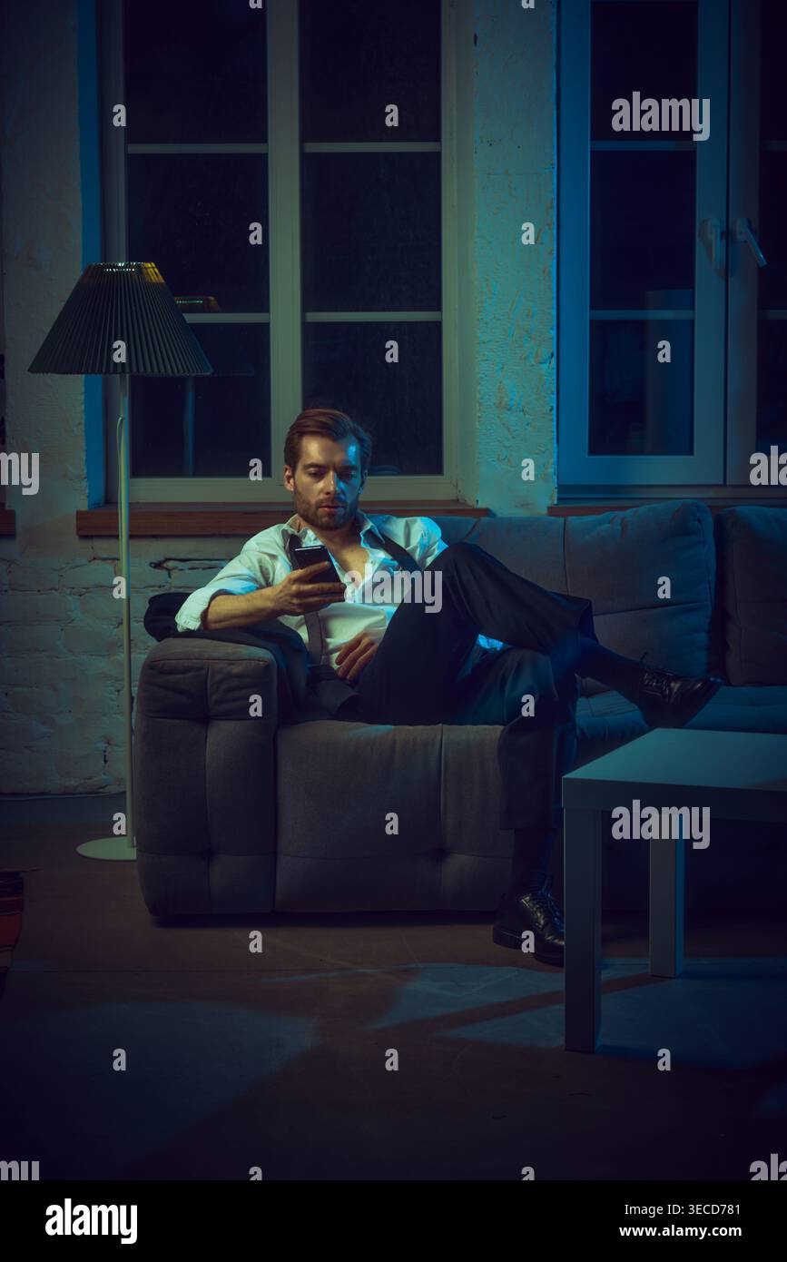 Man sitting on couch checking phone with serious expression in evening interior. Stock Photo