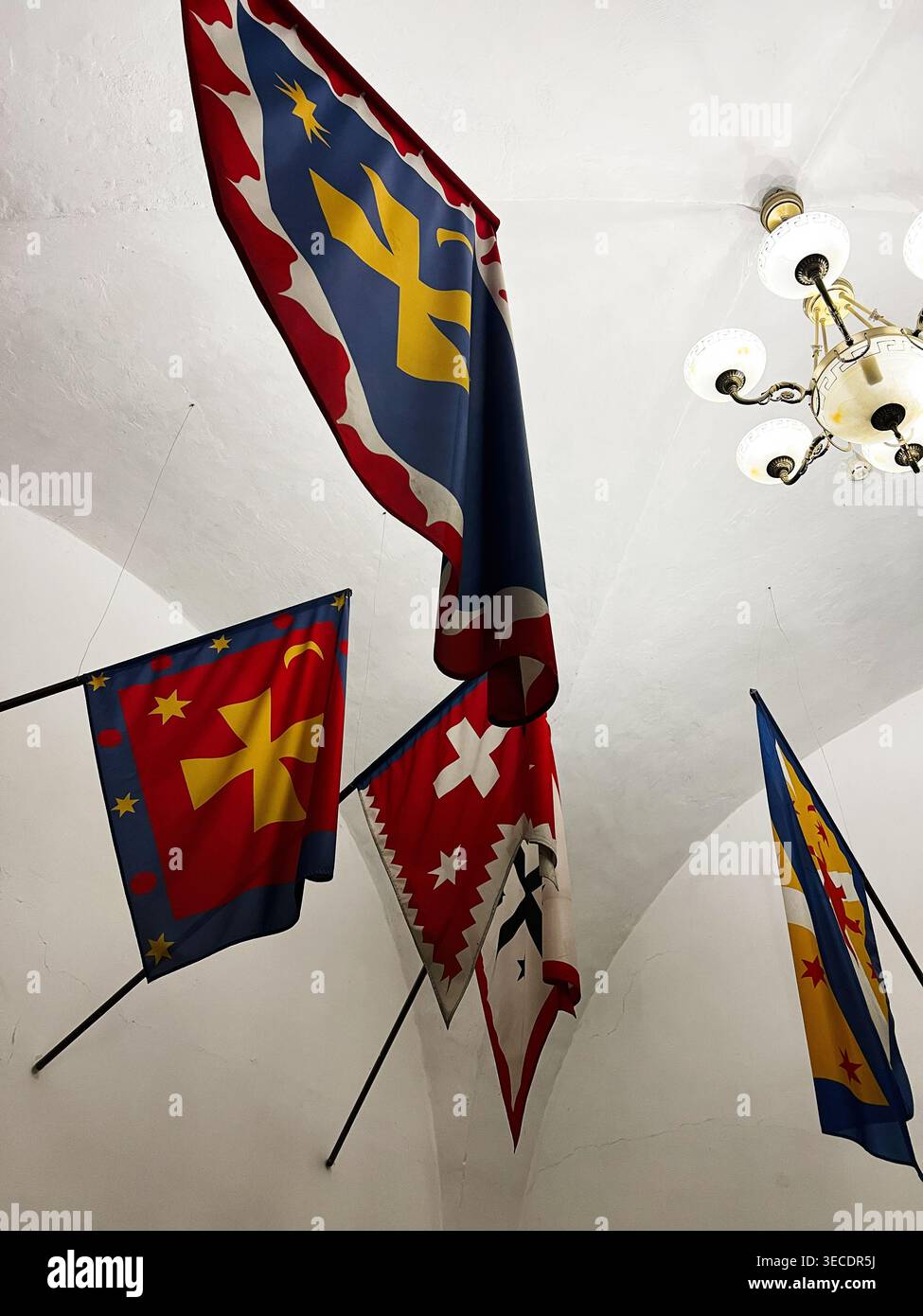 Zolochiv, Ukraine - April 28, 2026: Heraldic flags and banners with bright emblems hanging from a vaulted hall ceiling beside an ornate chandelier. Stock Photo