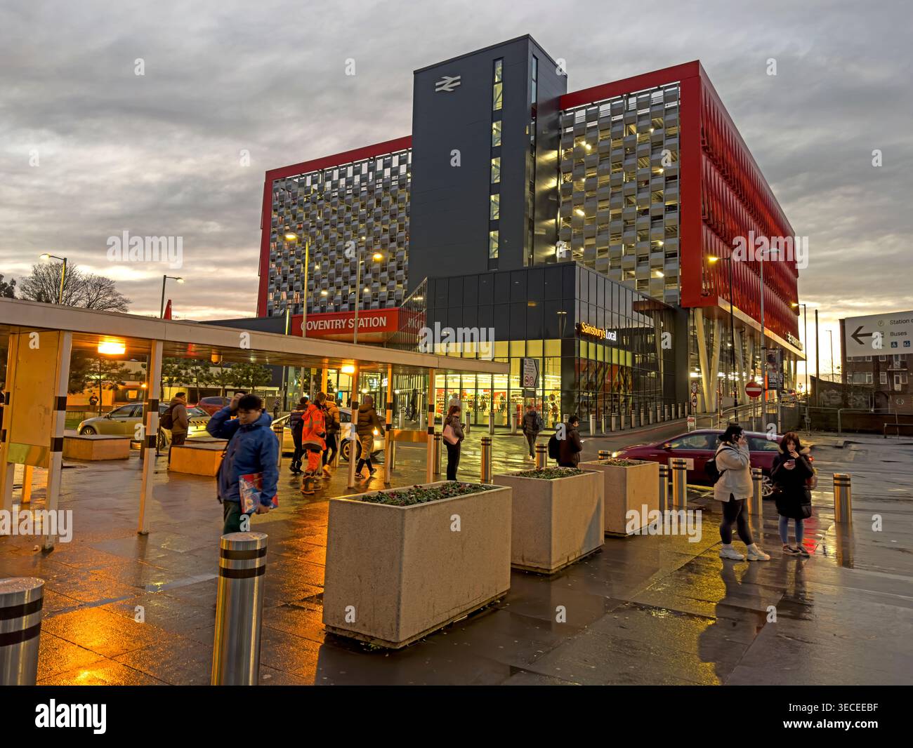 Coventry railway station at wet dusk, with passengers outside the modern red station frontage and Station Square entrance. Stock Photo