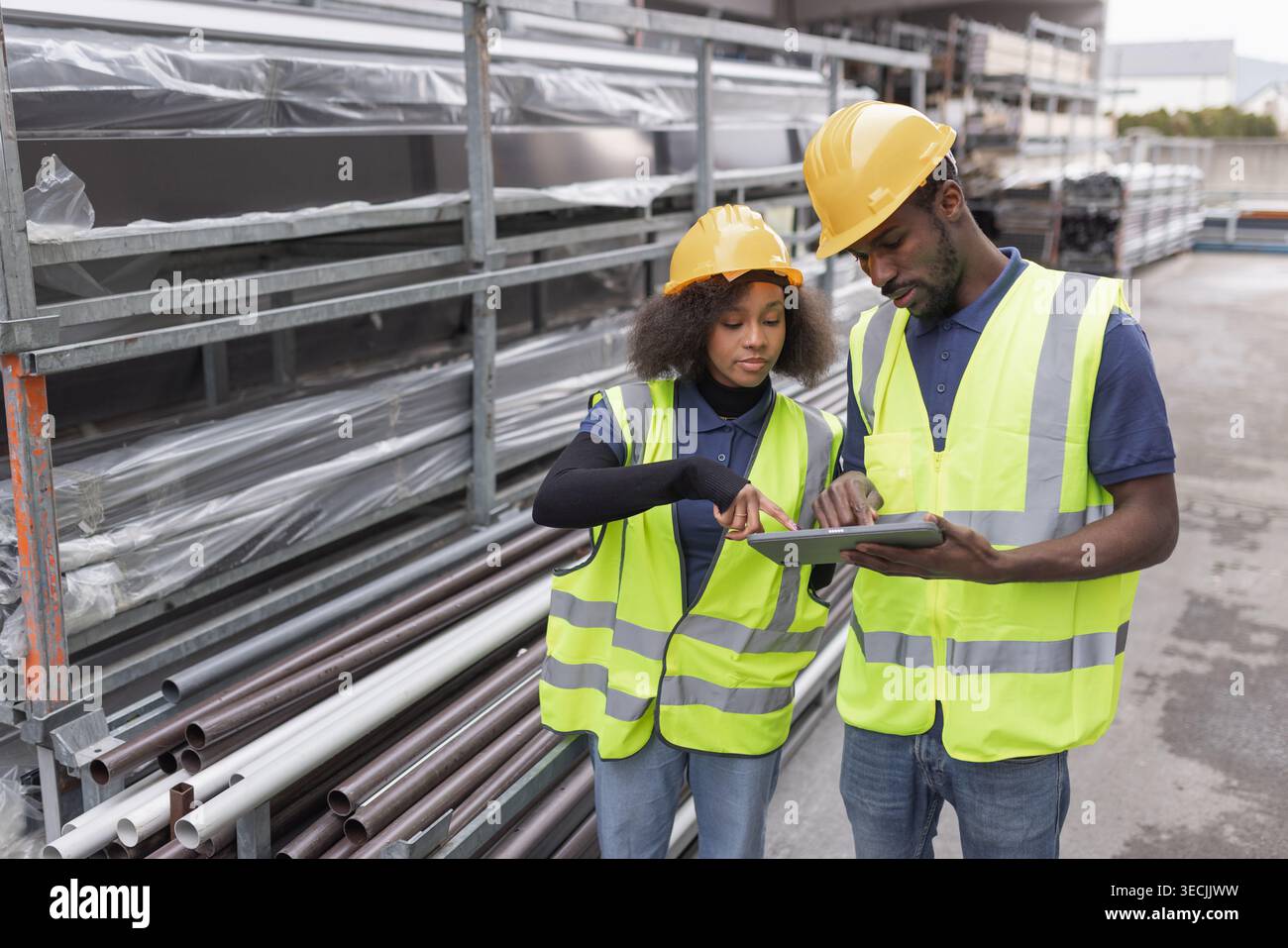 Two workers wearing protective gear, yellow hard hats and safety vests taking inventory in an outdoor warehouse using a digital tablet. Stock Photo