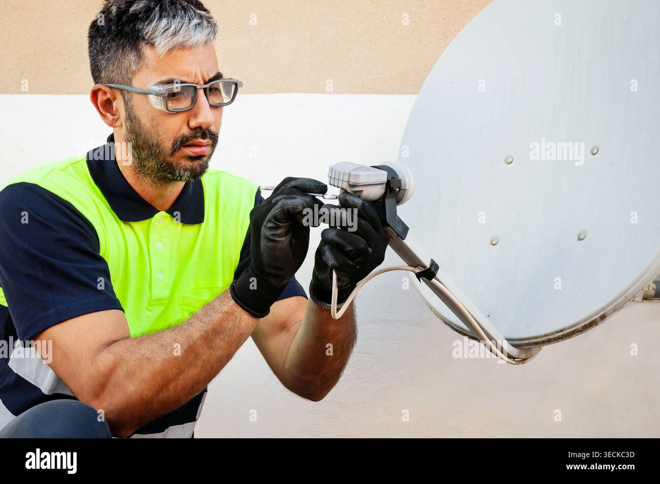 Technician wearing safety vest, gloves and eye protection performs maintenance on a satellite dish connection, ensuring occupational safety during out Stock Photo