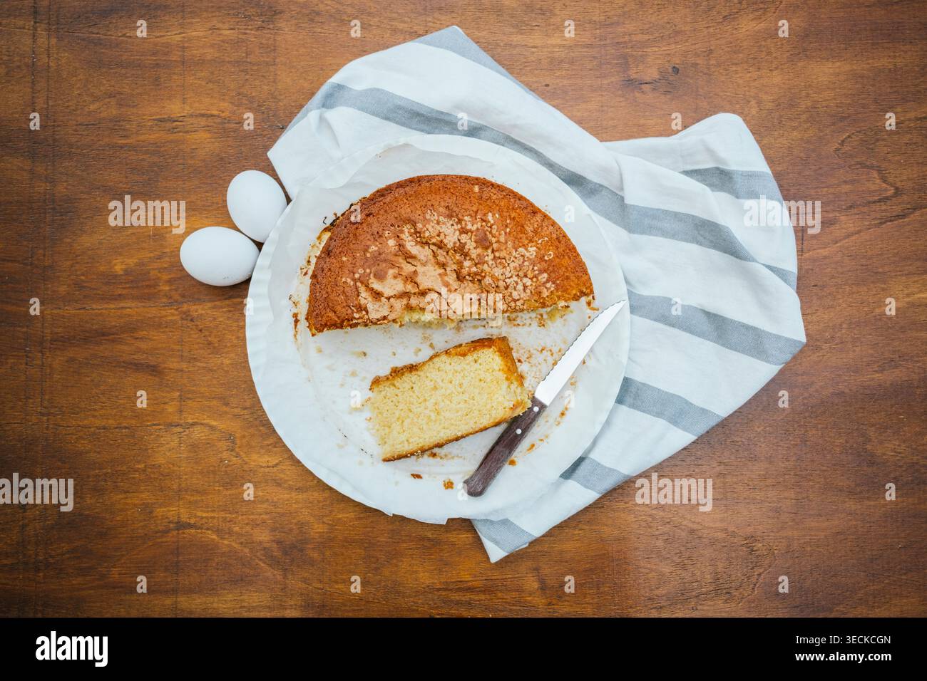 Overhead view of a homemade sponge cake with a slice cut out on a white plate, beside two eggs, a knife, and a striped towel on a rustic wooden table. Stock Photo
