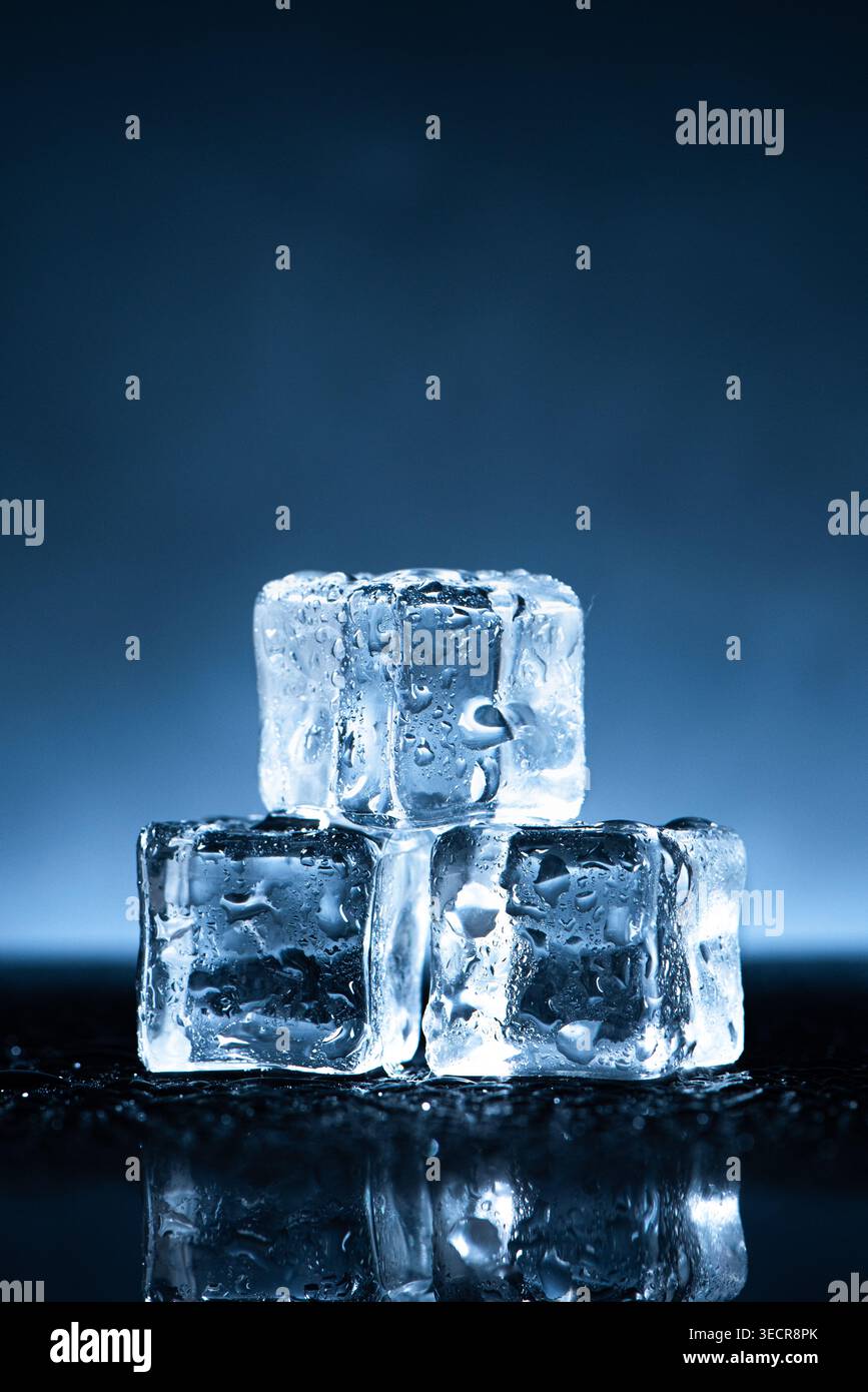 Three clear ice cubes with water drops reflecting on a dark surface Stock Photo