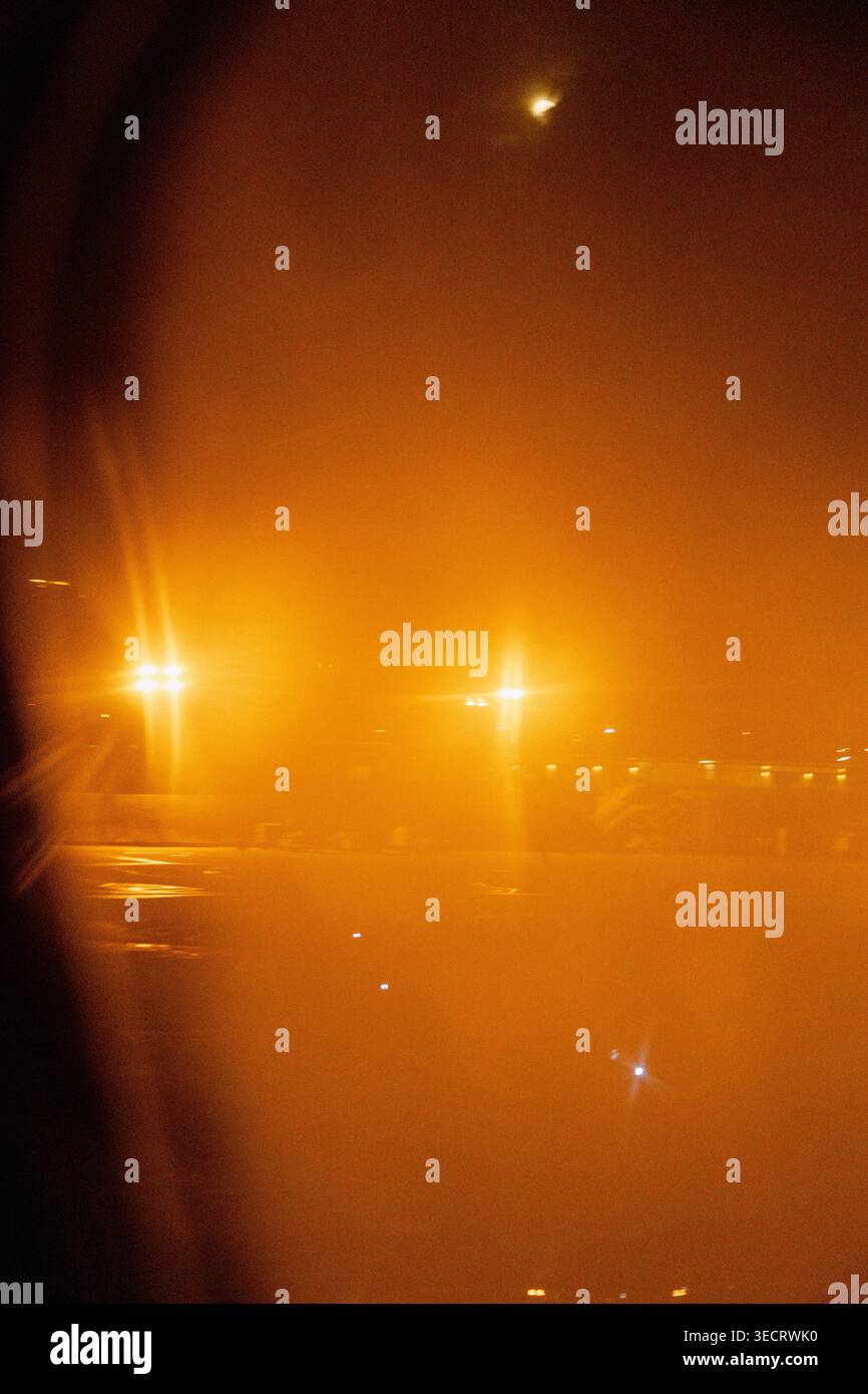 Nighttime view from an airplane window shows a foggy airport apron with glowing runway lights and reflections on wet pavement, creating a warm, hazy t Stock Photo