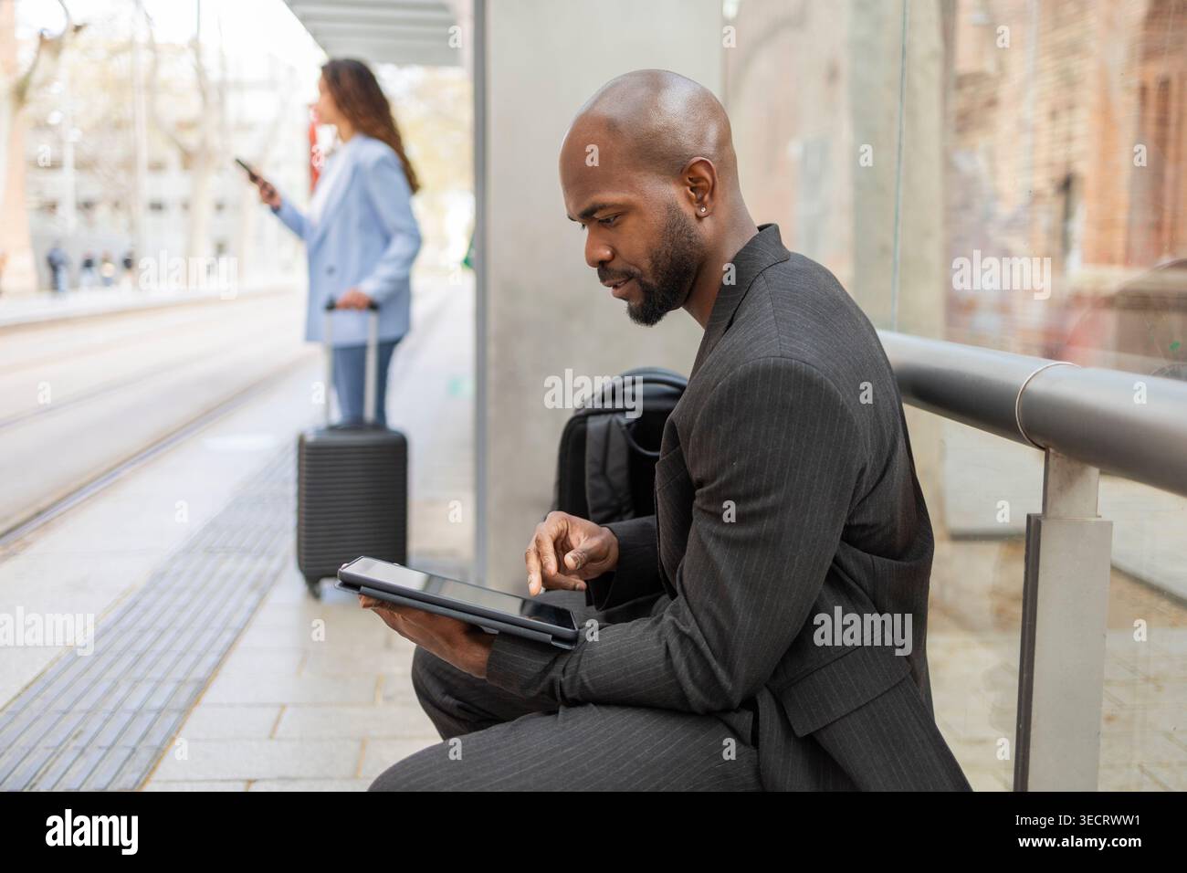 Focused businessman in suit using a tablet at a transit stop while colleague with suitcase checks phone in background, capturing business mobility and Stock Photo