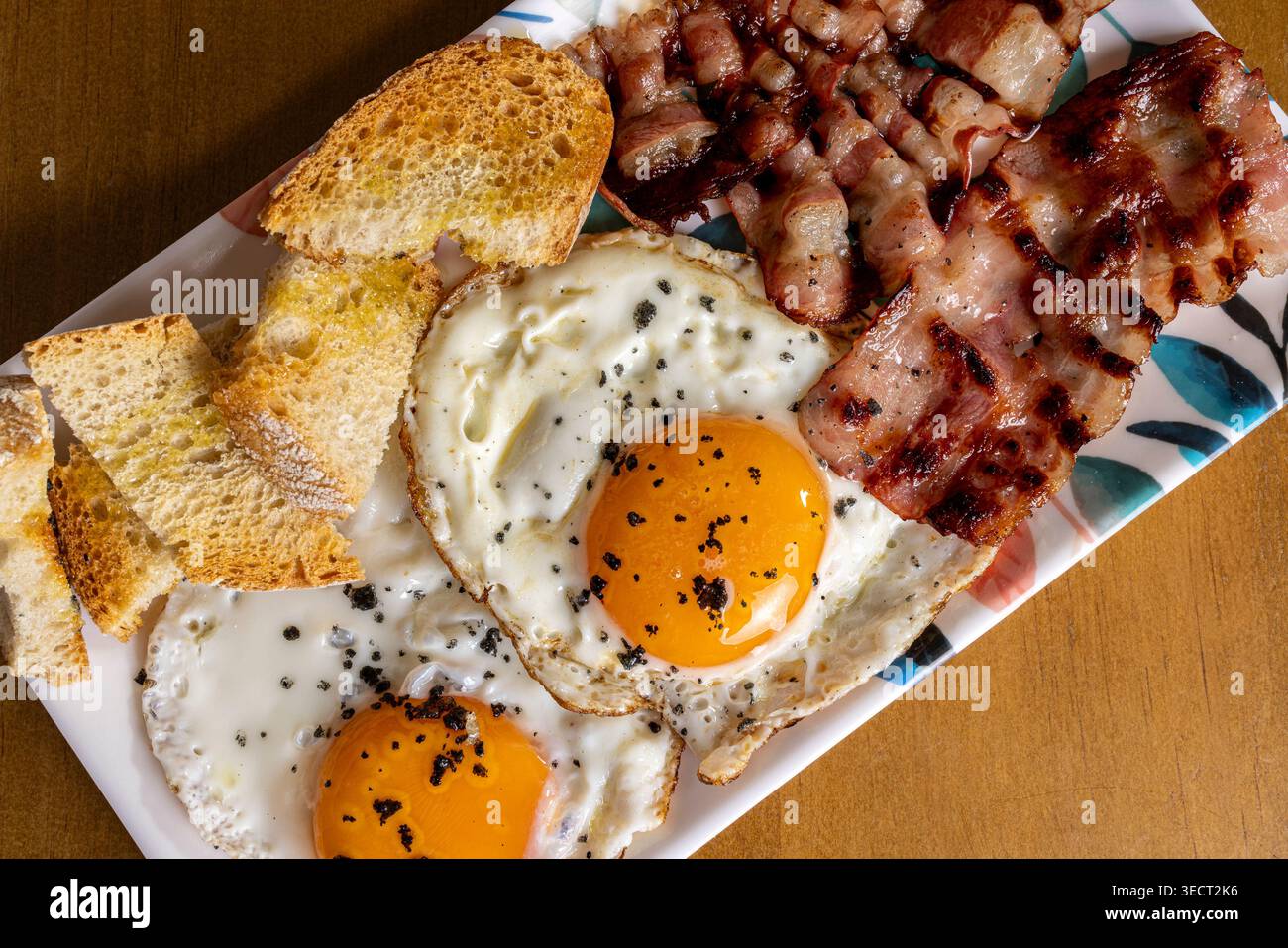 Close-up of a hearty morning breakfast plate with two sunny-side fried eggs, crispy bacon strips, and sliced rustic bread on a patterned dish, ready t Stock Photo