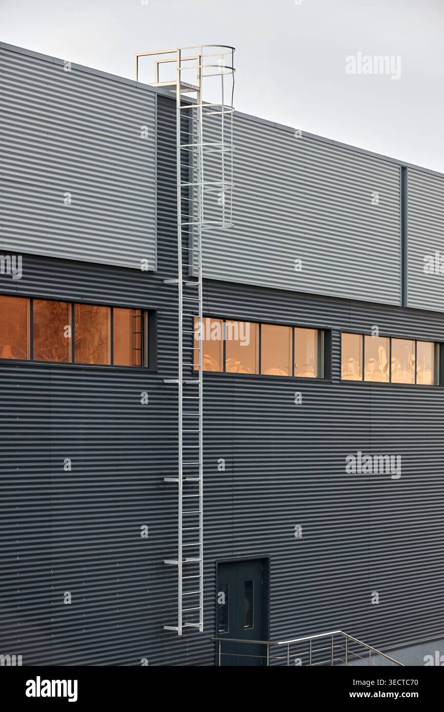 Architectural facade with orange cladding corrugated metal panels, industrial vertical ladder with a safety cage or crinoline, large glass window. Bui Stock Photo