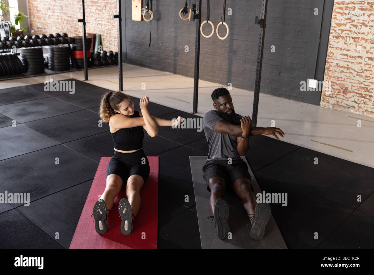 Diverse exercise partners stretching arms while sitting on red and dark mats at gym with rings Stock Photo