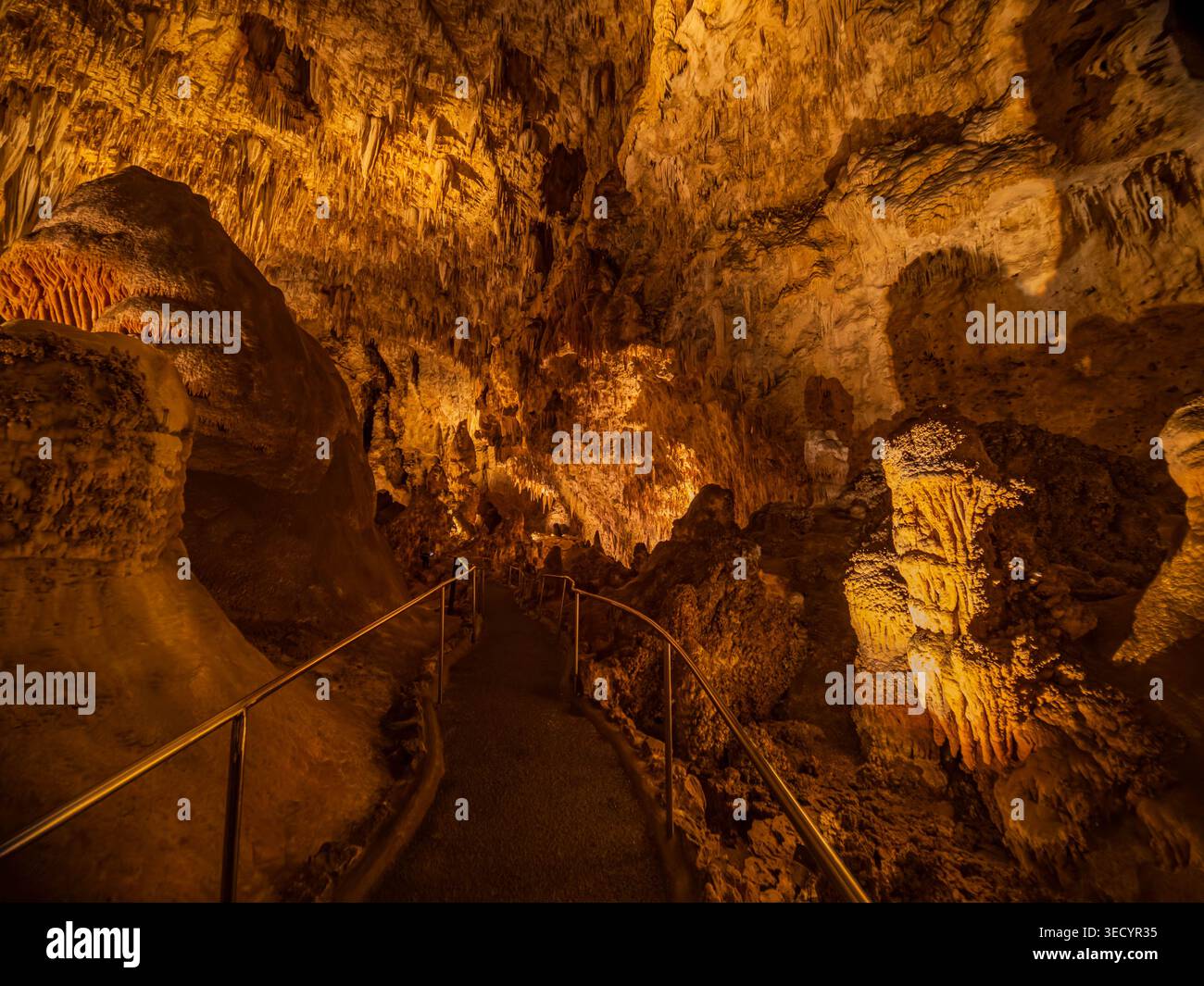 A paved path winds between towering rock formations and stalactites illuminated by warm orange lighting in a deep cavern. Stock Photo