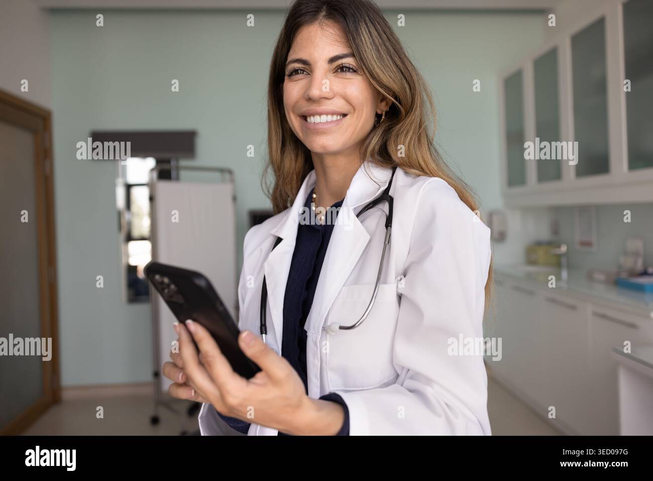 Smiling Latina woman doctor in white lab coat using smartphone Stock Photo